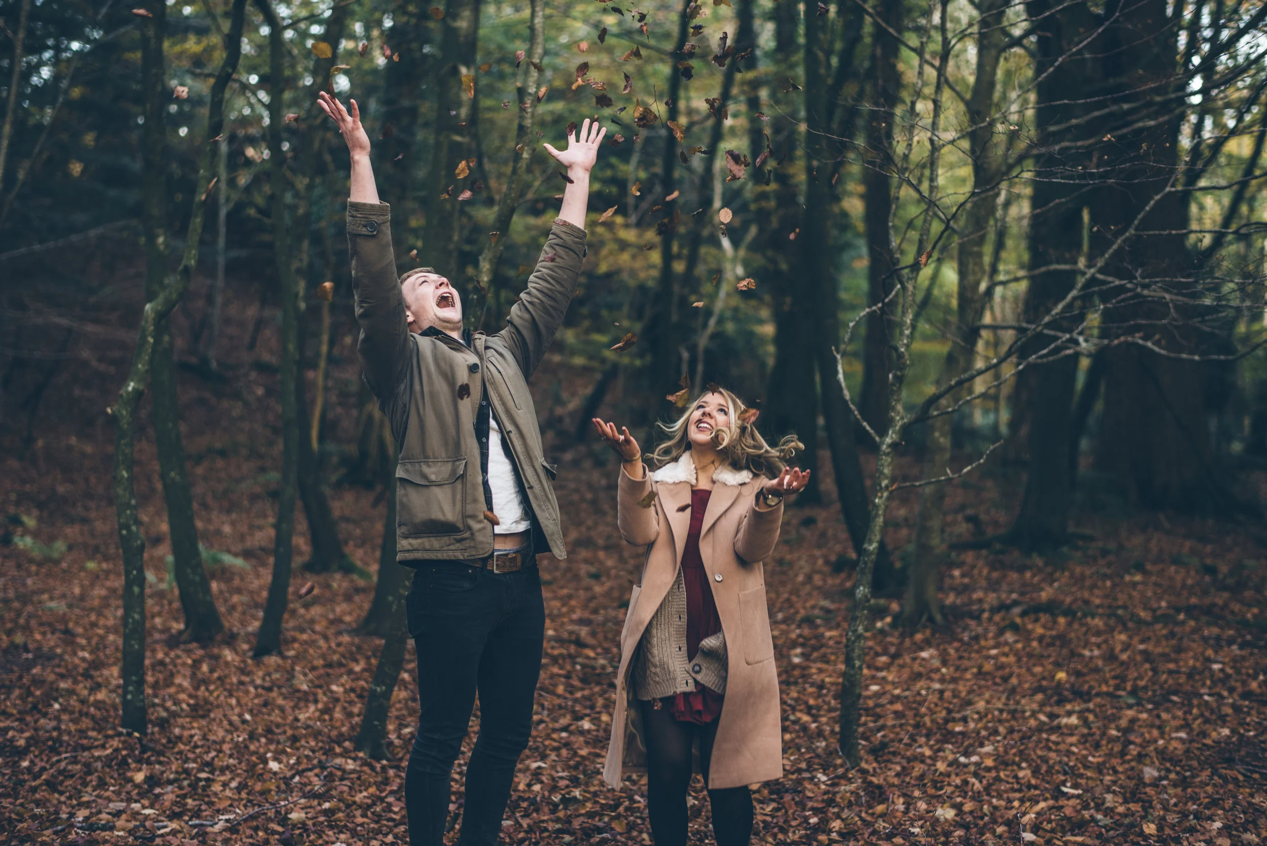 A young man and woman standing in a forest, smiling and throwing leaves into the air.