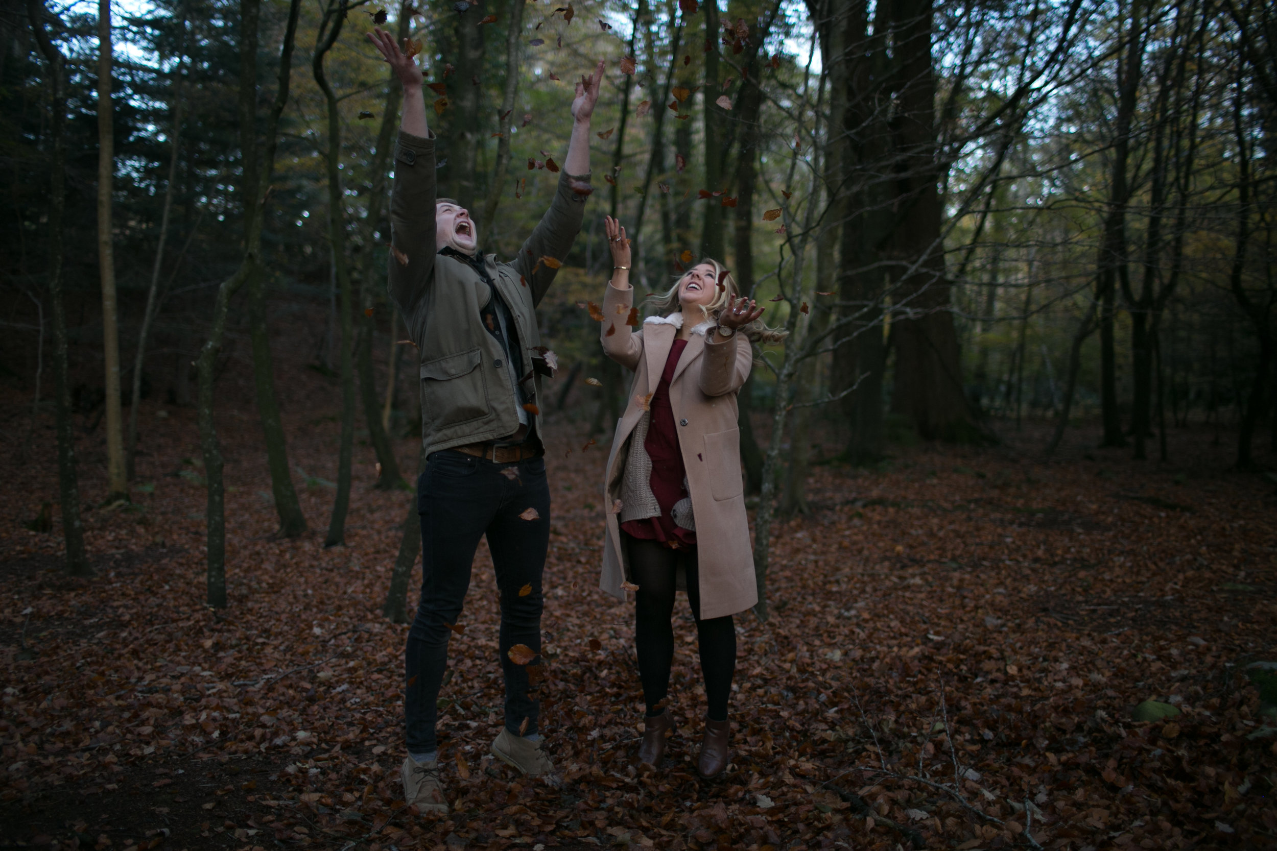 Two people standing in a wooded area, joyfully throwing fallen leaves into the air with trees and a forest floor covered in leaves in the background.