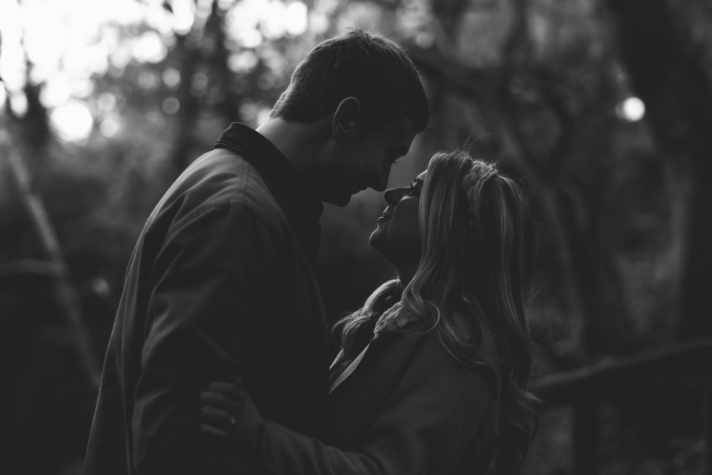 A black and white photo of a couple standing close together, touching foreheads and smiling, outdoors with blurred trees in the background.