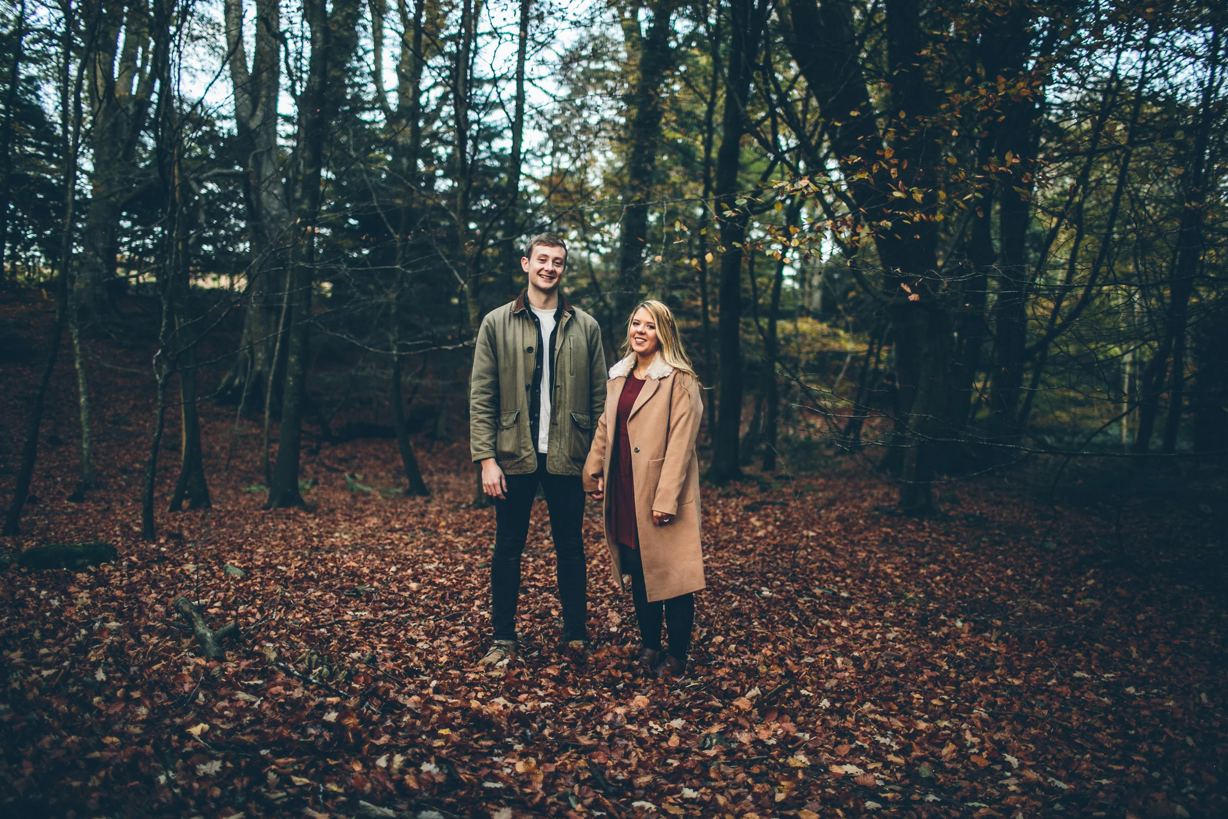 A young man and woman standing hand in hand in a forest with fallen autumn leaves, trees with sparse foliage in the background.