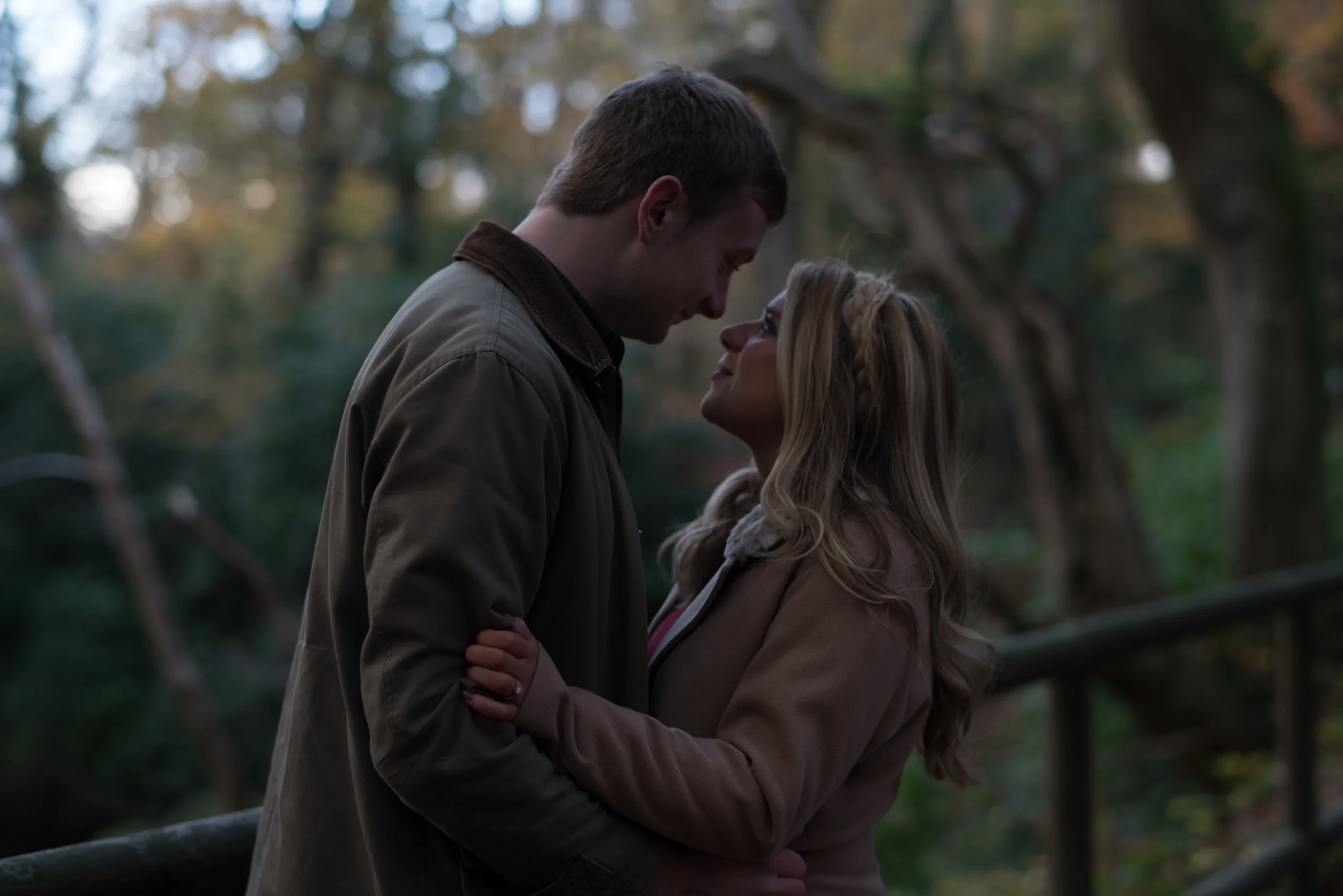 A couple standing close together outdoors, looking into each other's eyes, surrounded by trees in a wooded area during daytime.