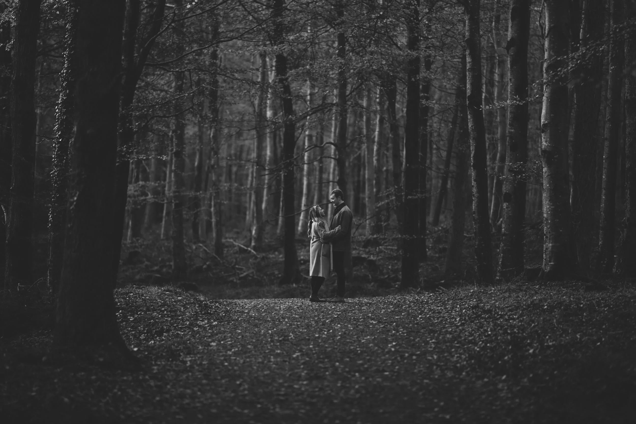 A black and white photo of a couple standing closely together in a dense forest, looking at each other.