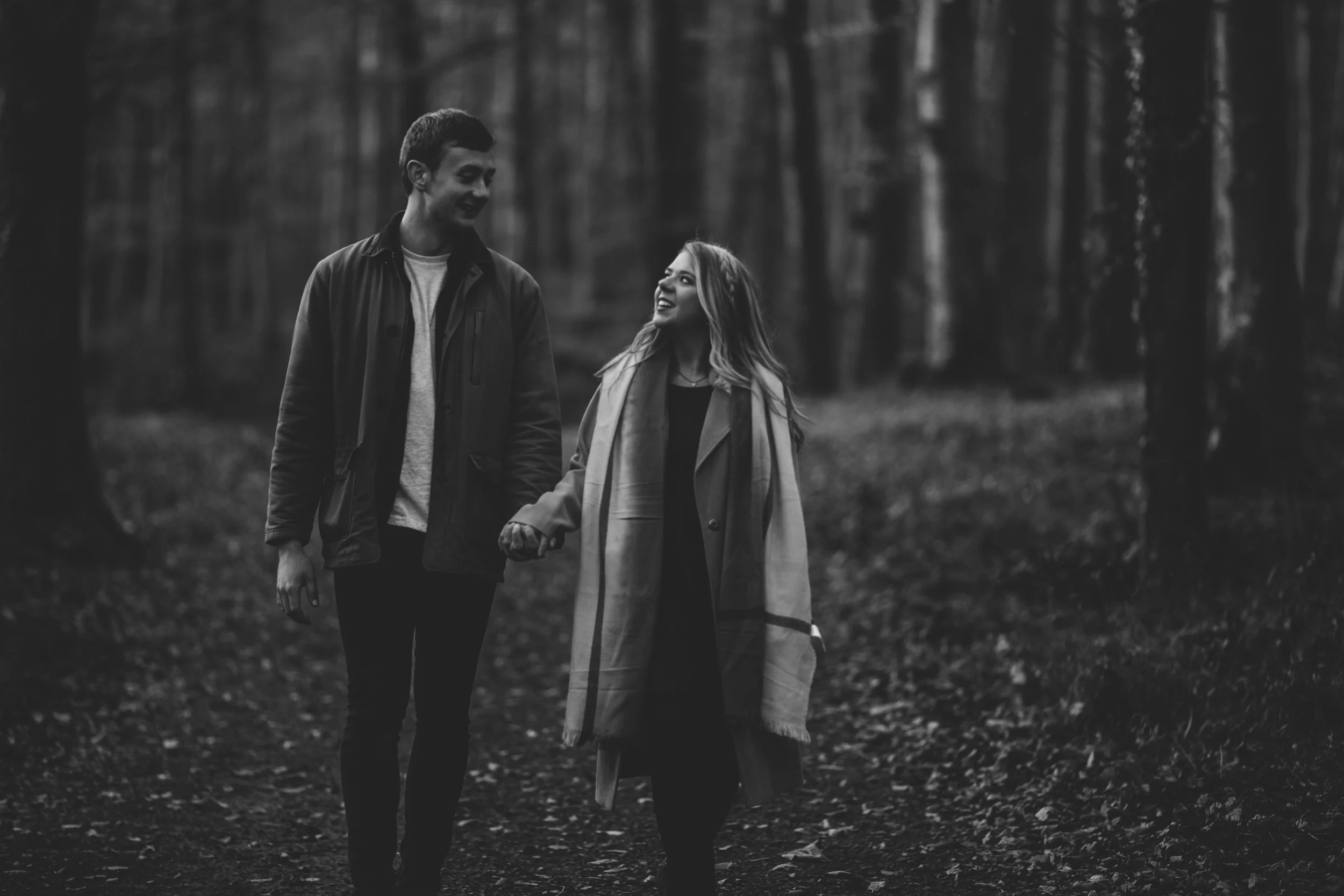 A young couple walking hand in hand through a forest, smiling and looking at each other.