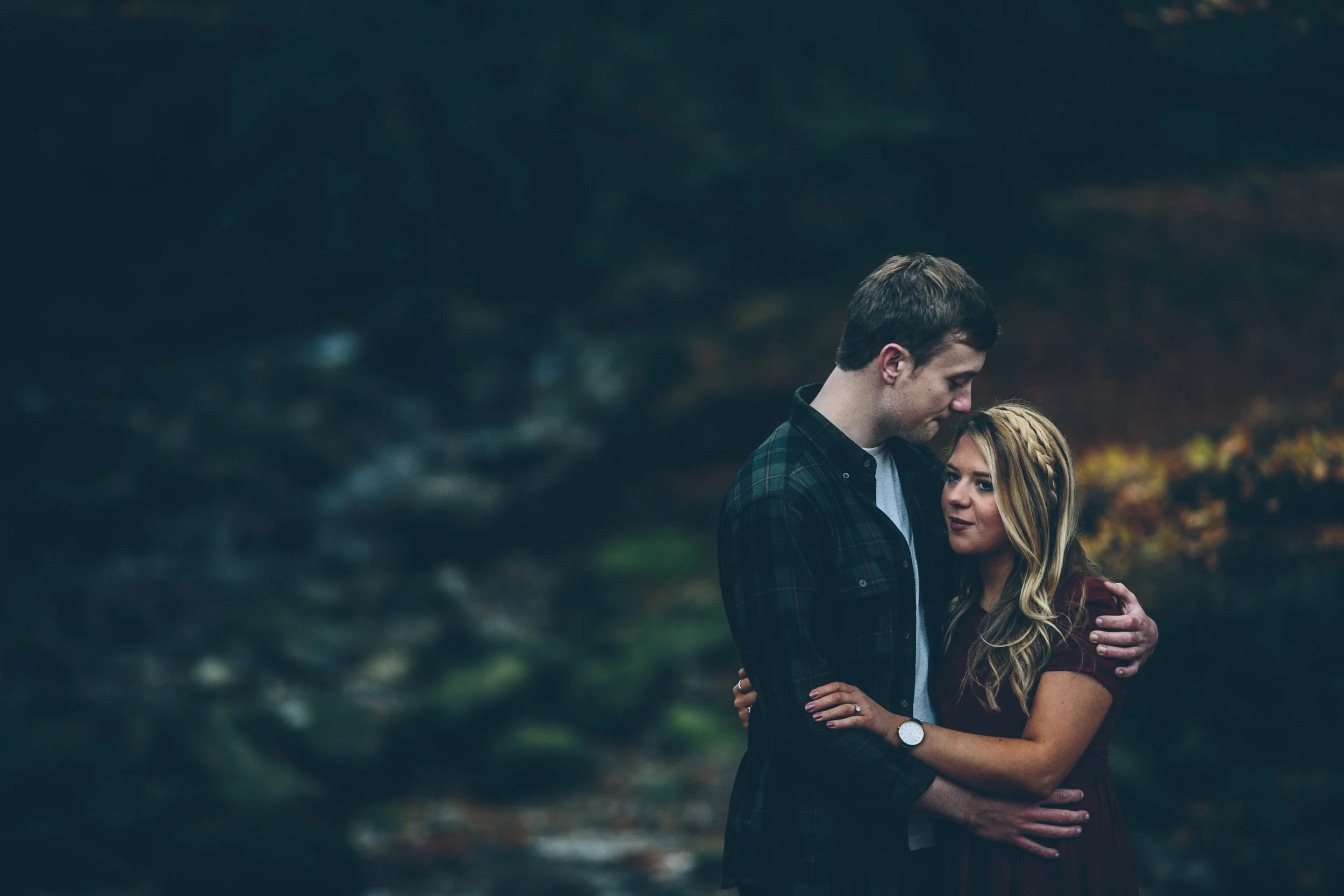 A young couple embracing outdoors in a dark, wooded area with a stream in the background.