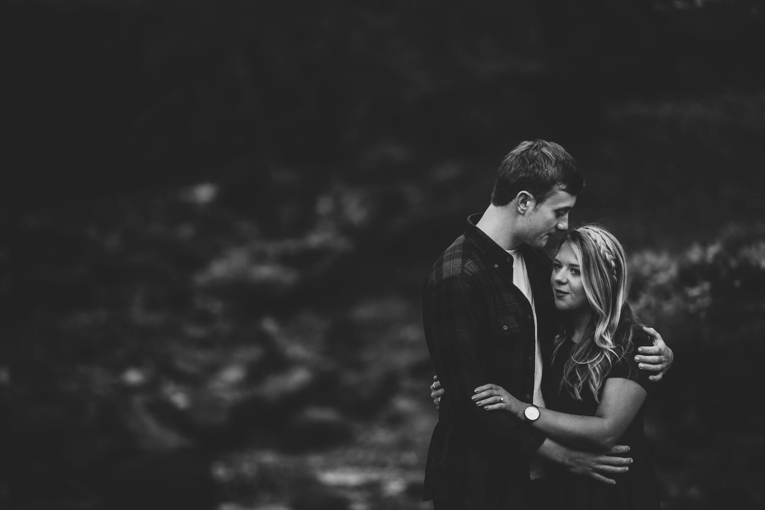A black and white photo of a couple embracing outdoors, with the man gently touching the woman's head, both looking at each other.