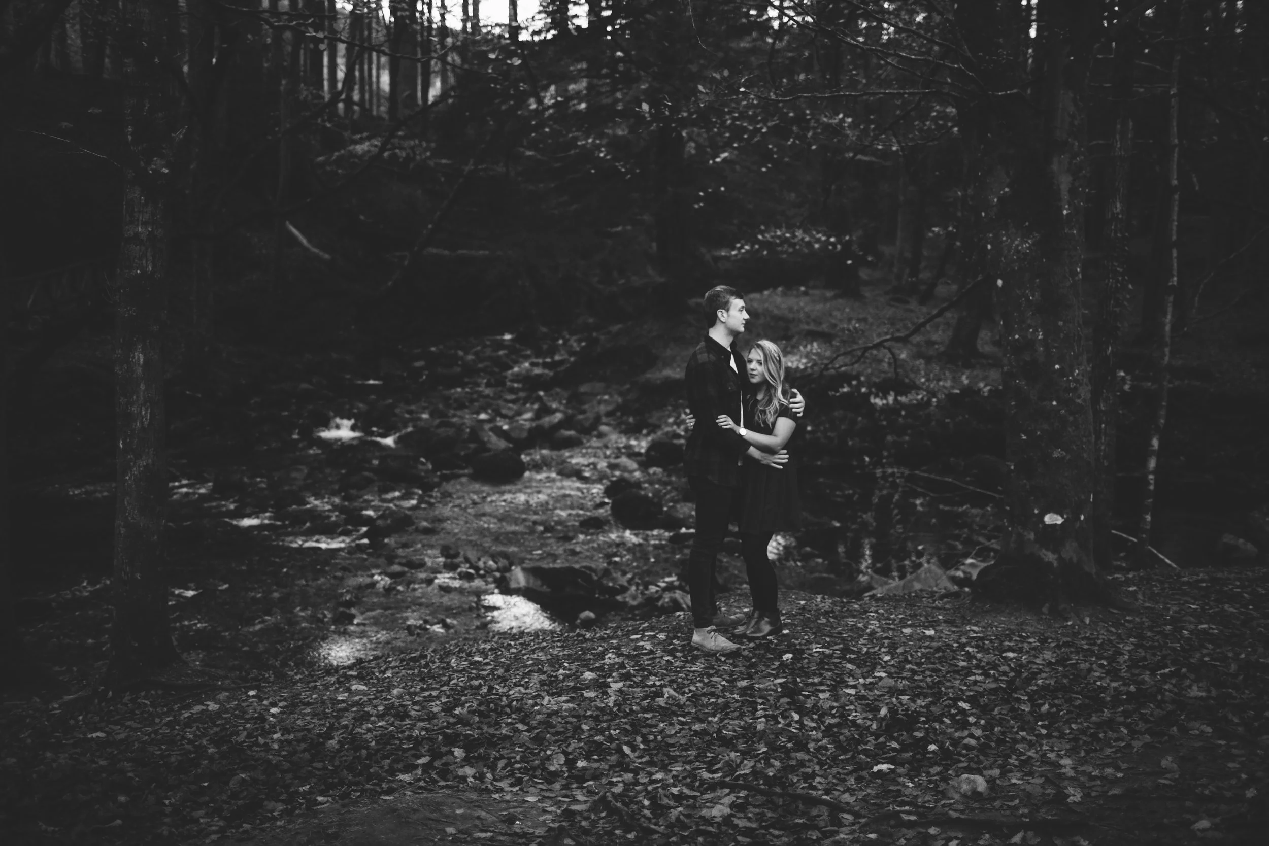 A black and white photo of a young man and woman embracing in a forest by a small stream.
