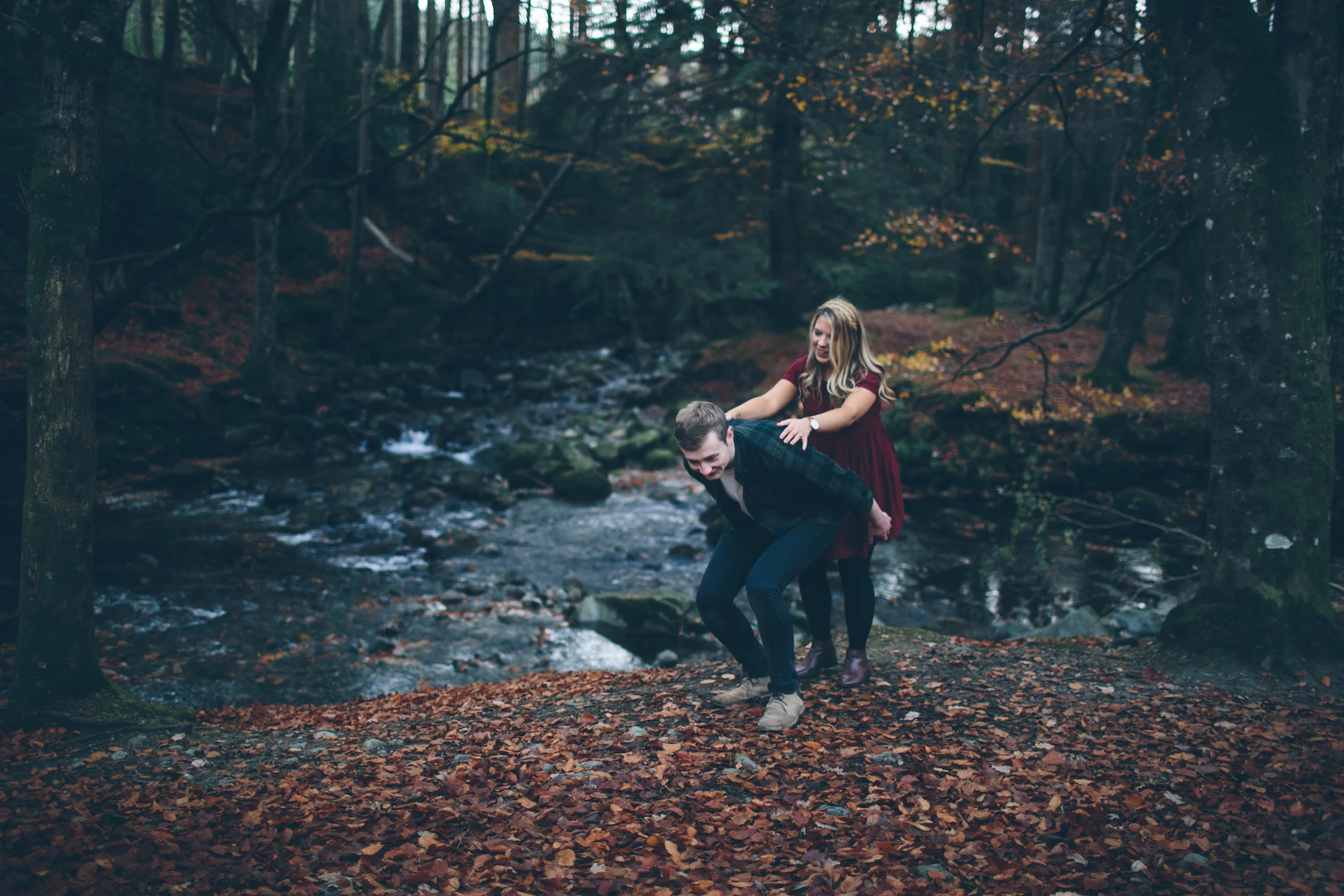 Two young adults, a man and a woman, playfully wrestling on a leaf-covered forest ground near a creek, surrounded by trees with autumn foliage.