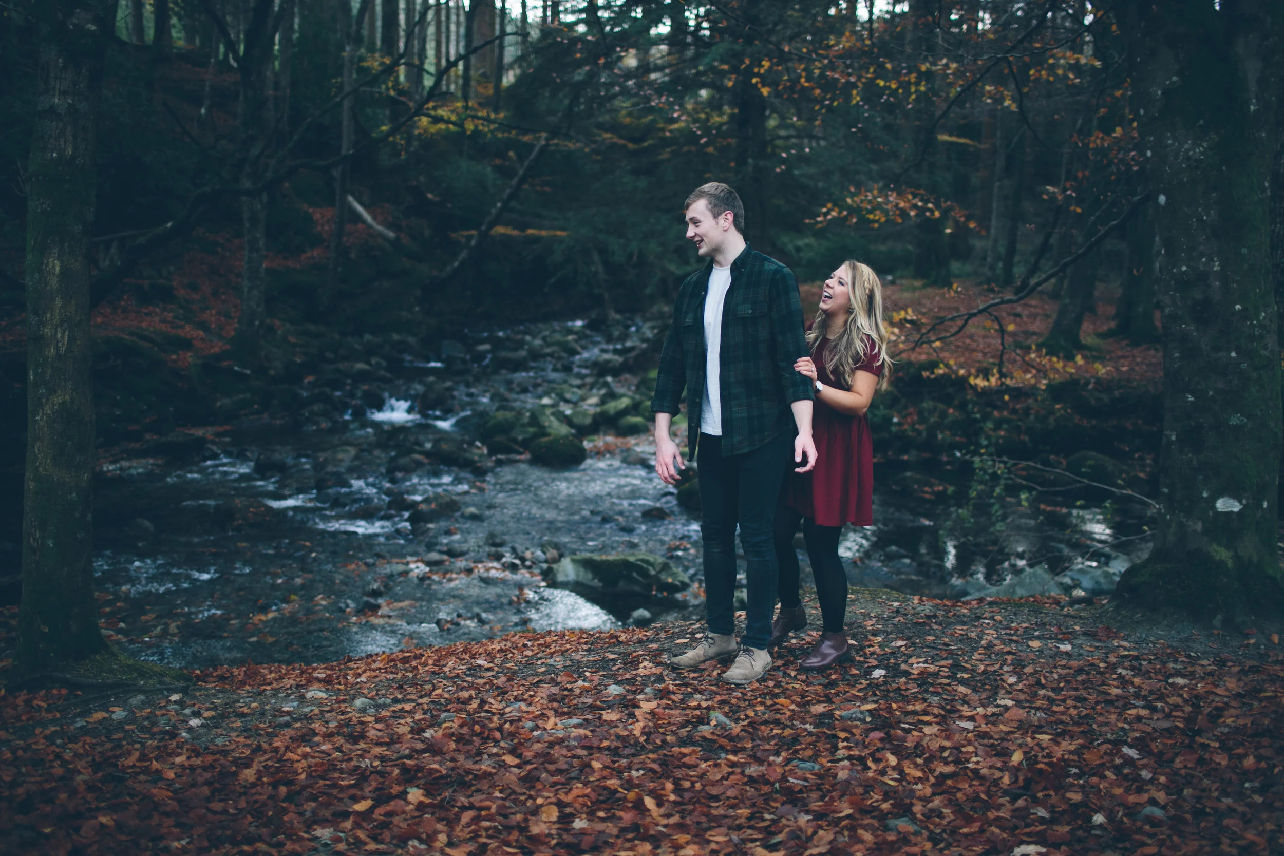 A young couple walking along a leaf-covered trail next to a stream in a forest during autumn, smiling and enjoying each other's company.