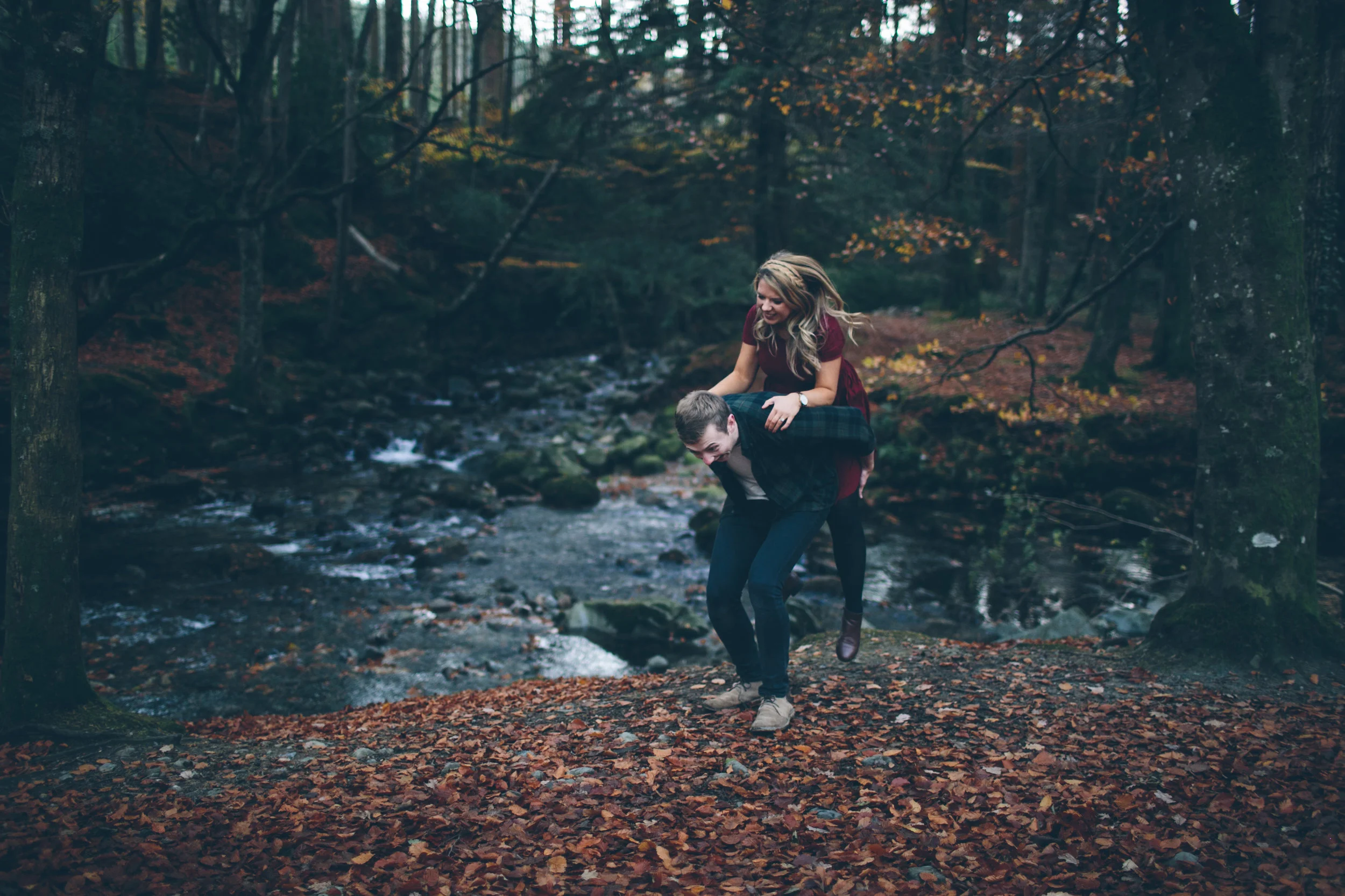 Two people, a man and a woman, playing around near a stream in a forest during autumn, with fallen leaves covering the ground.