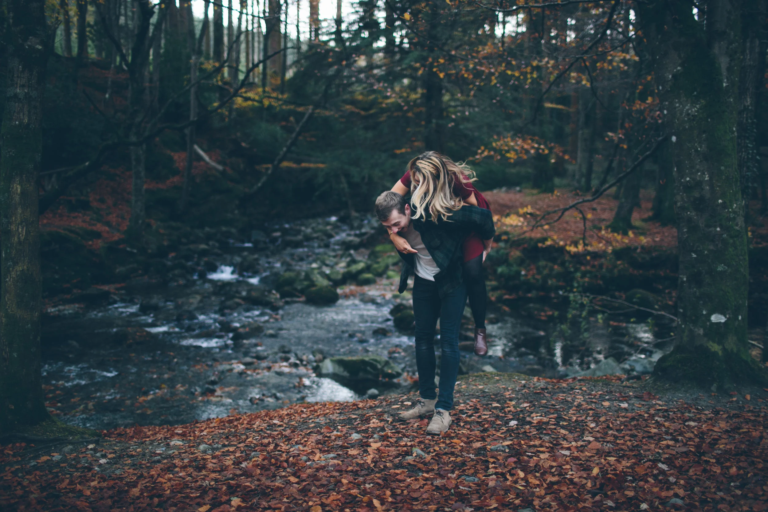 A couple in an autumn forest, with the man giving the woman a piggyback ride over fallen leaves near a stream.