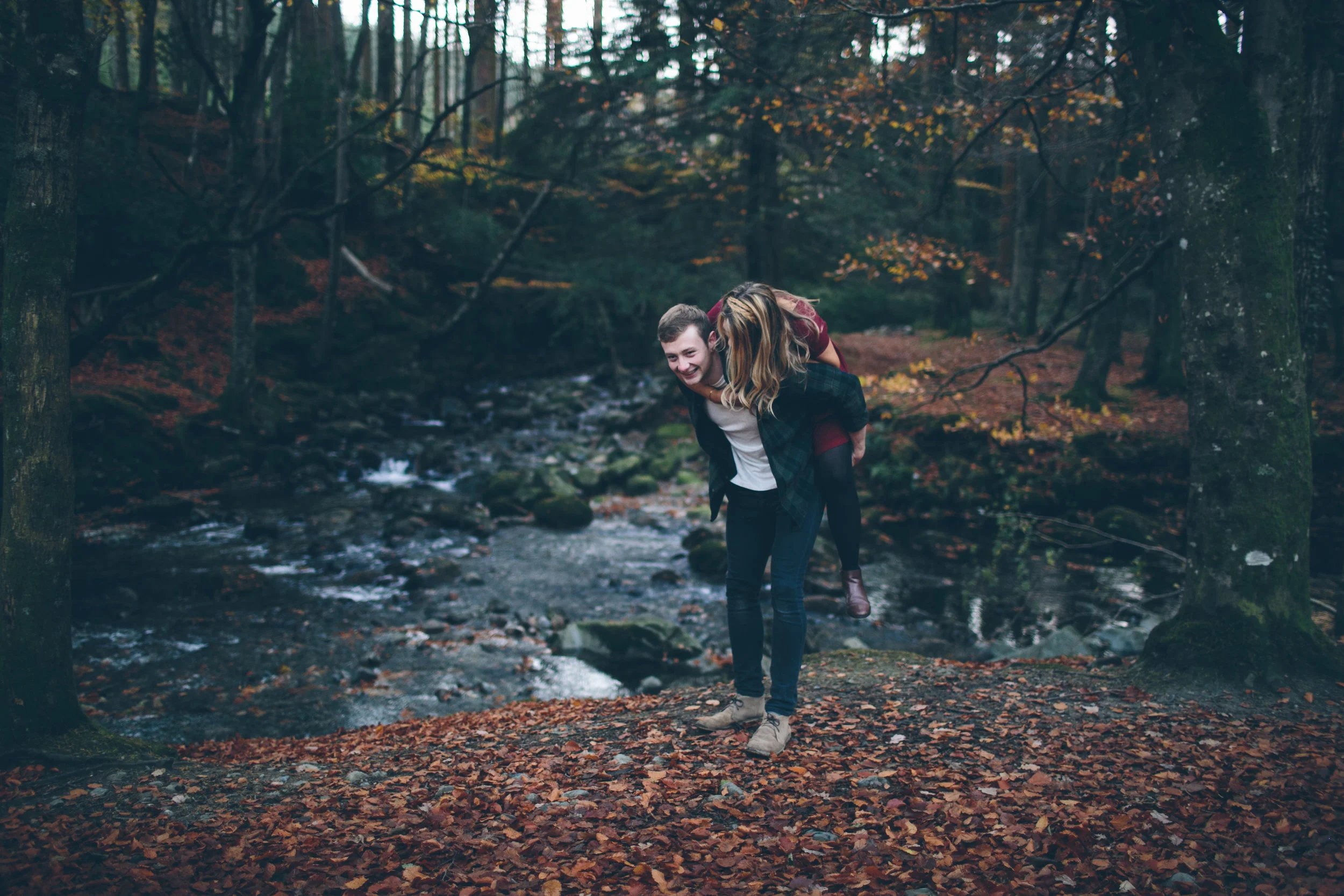 A young man giving a piggyback ride to a young woman on a forest trail beside a creek with autumn foliage.