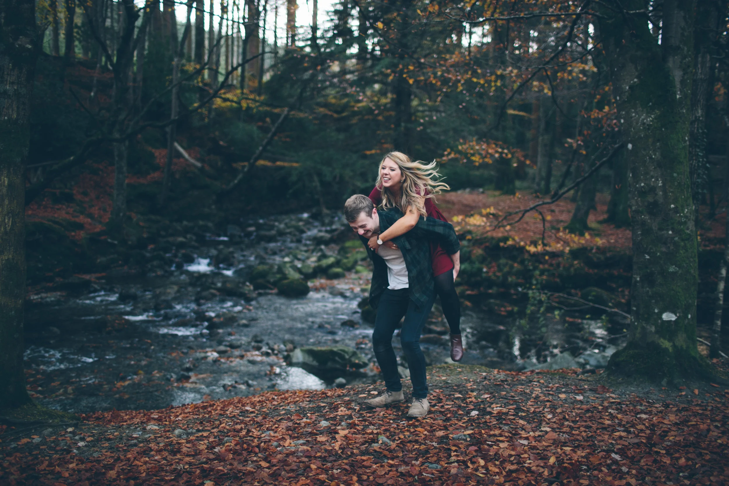 A young couple enjoying a piggyback ride by a stream in a forest with autumn leaves.