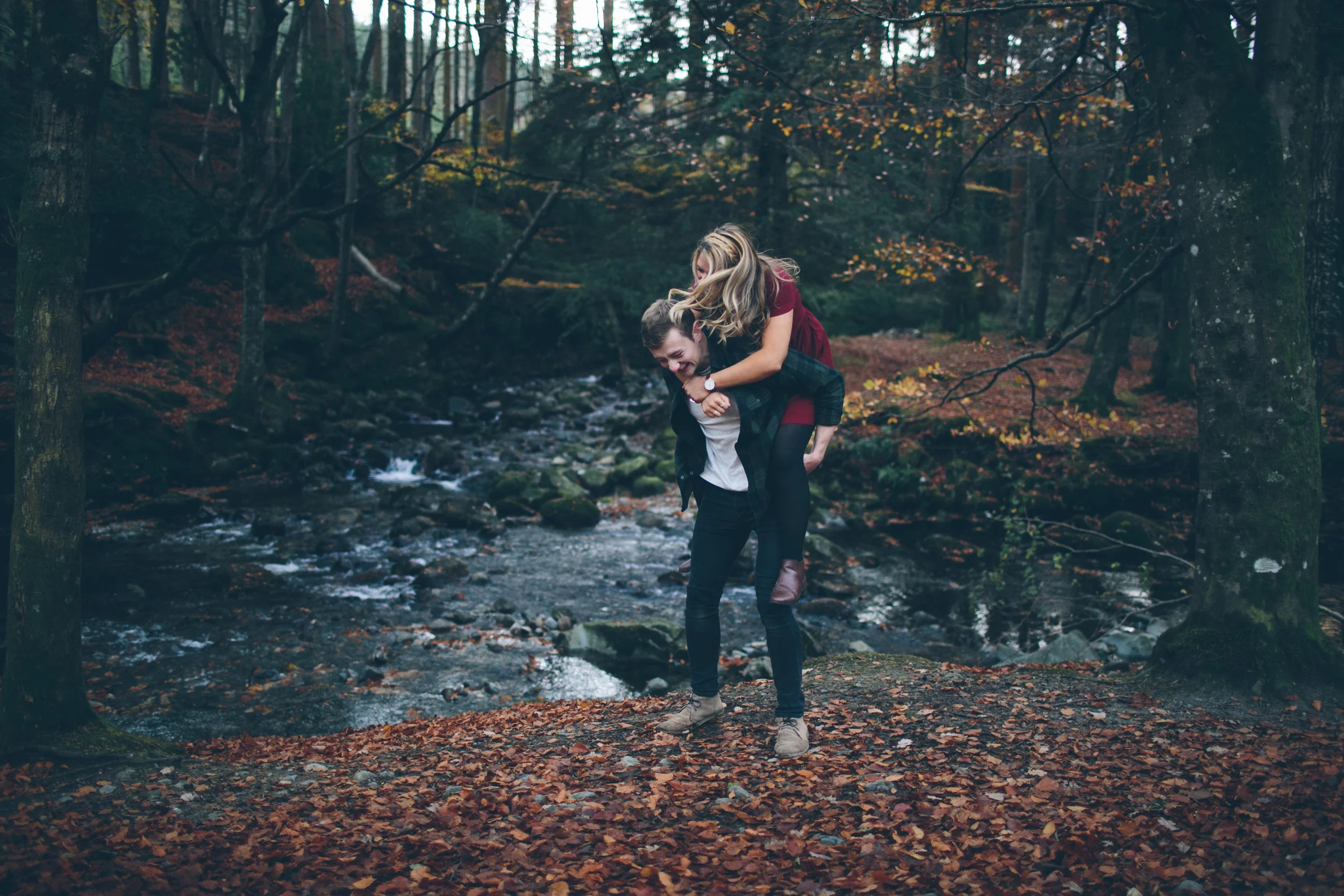 A couple is enjoying an outdoor moment in the woods, with the woman giving the man a piggyback ride by a stream surrounded by autumn leaves.