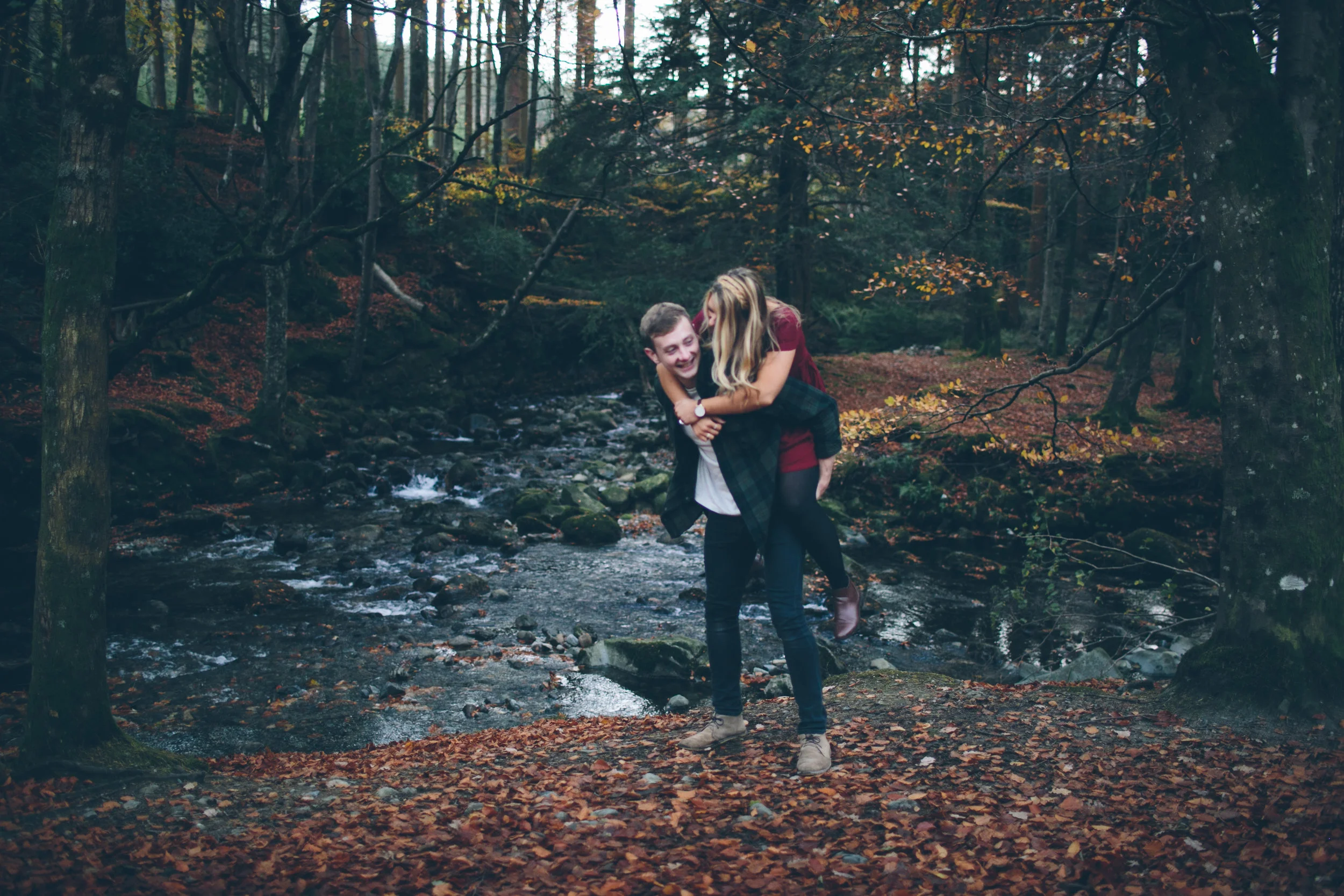 A couple smiling and laughing, with the woman giving a piggyback ride to the man while standing near a creek in a forest during autumn.