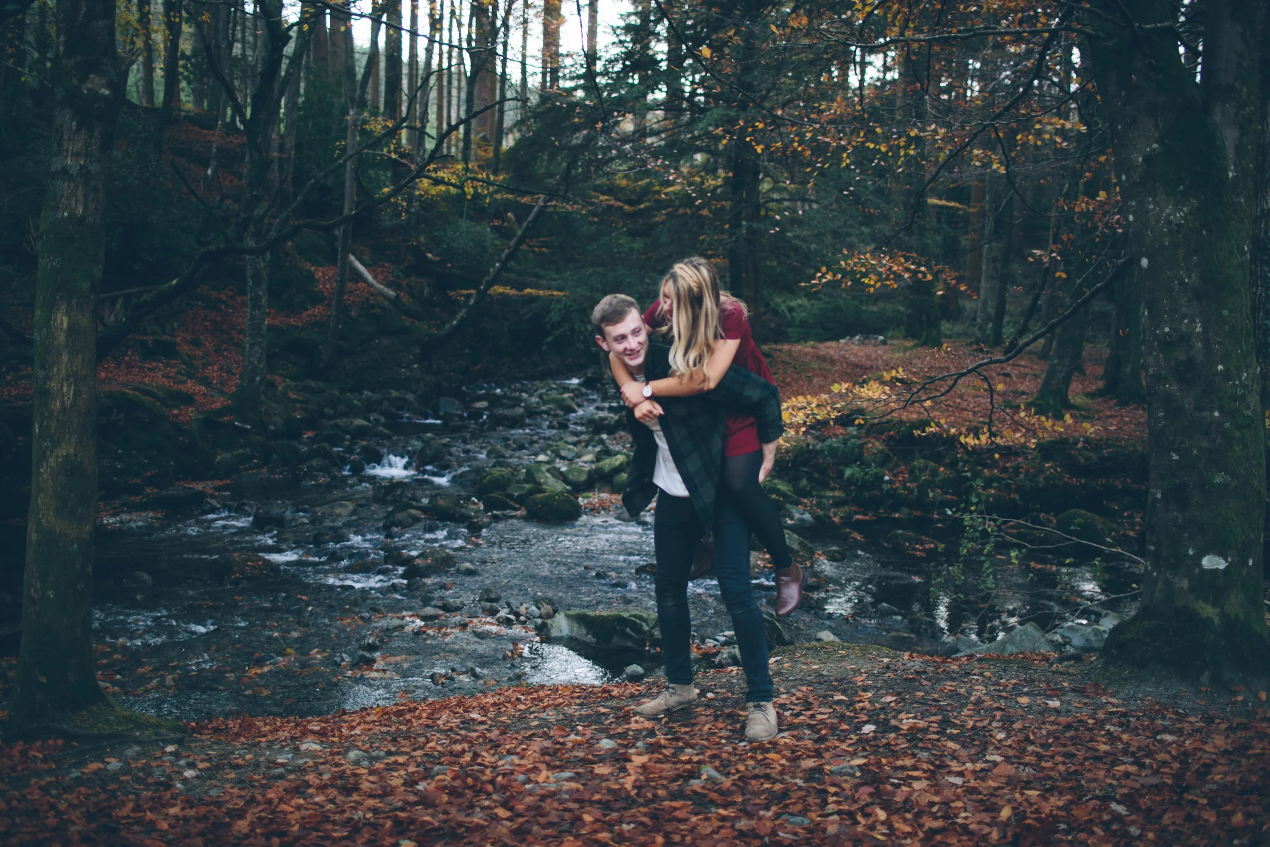 A young couple in a forest by a stream, with the man piggybacking the woman, both smiling and enjoying each other's company during autumn.