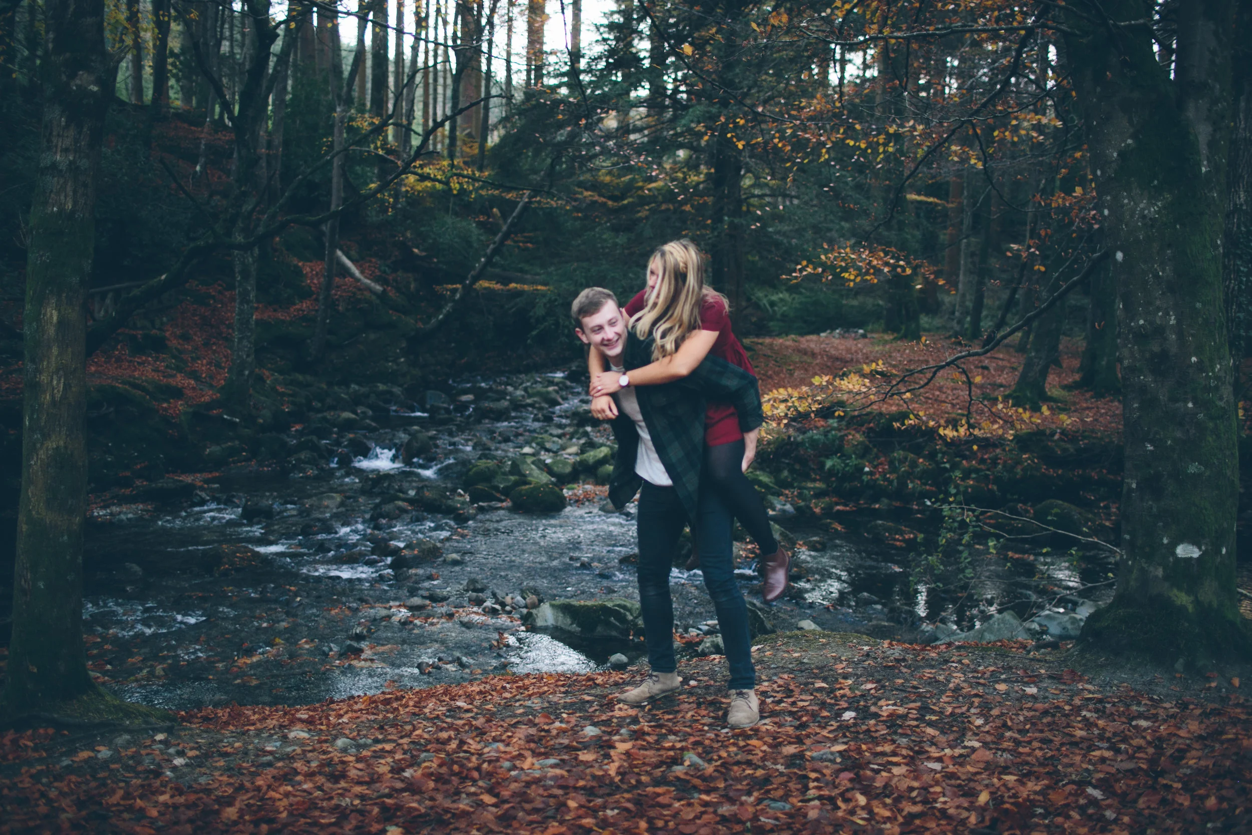 A young man giving a piggyback ride to a young woman in a wooded area with a stream, surrounded by autumn leaves.