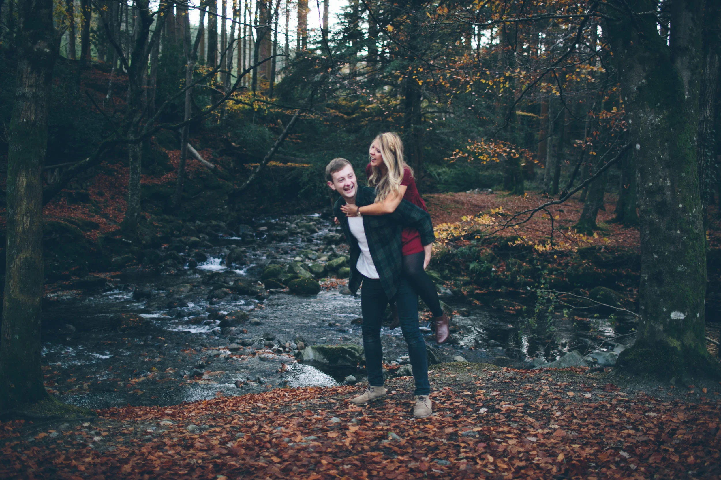 A young man with a young woman on his back standing on fallen autumn leaves near a creek in a forest, both smiling and laughing.