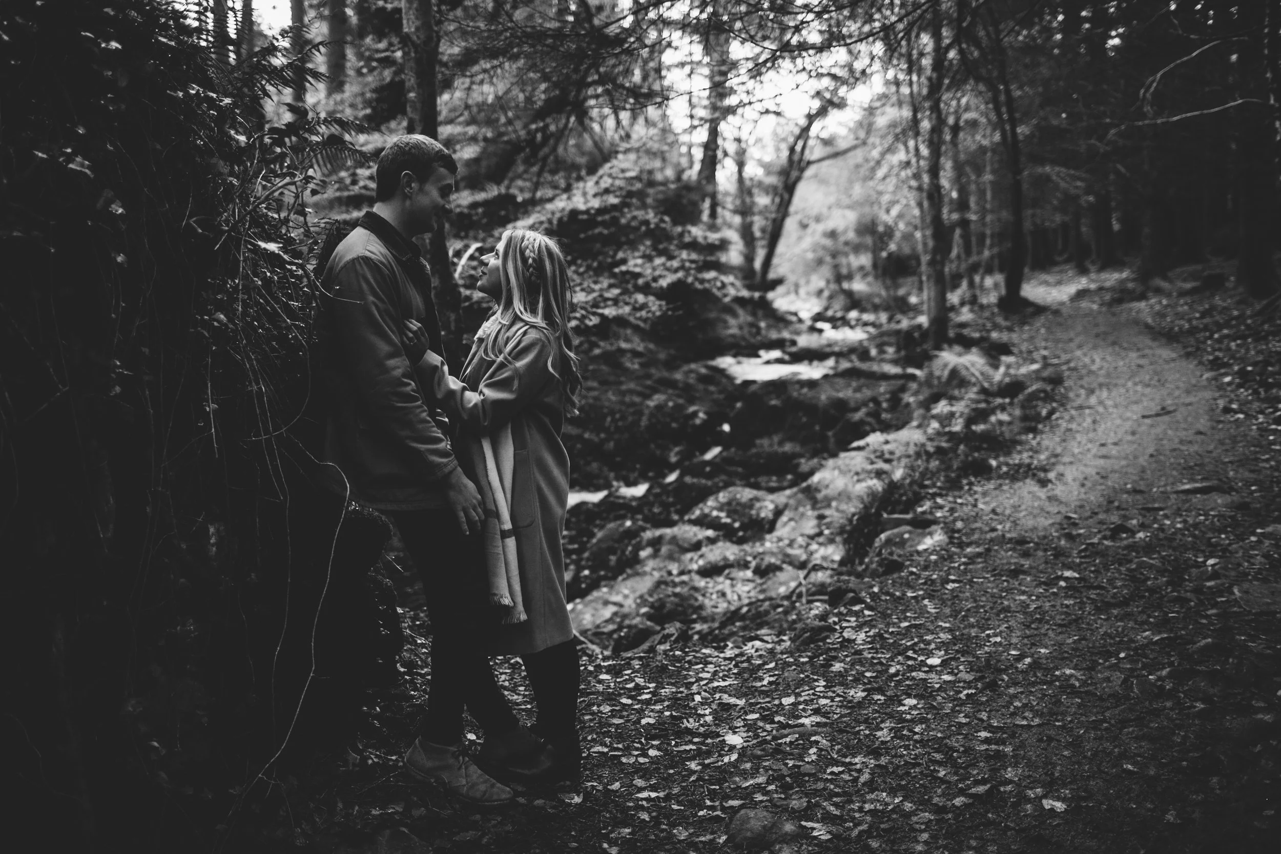 A black and white photo of a man and woman standing close together on a nature trail in a forest, gazing into each other's eyes.