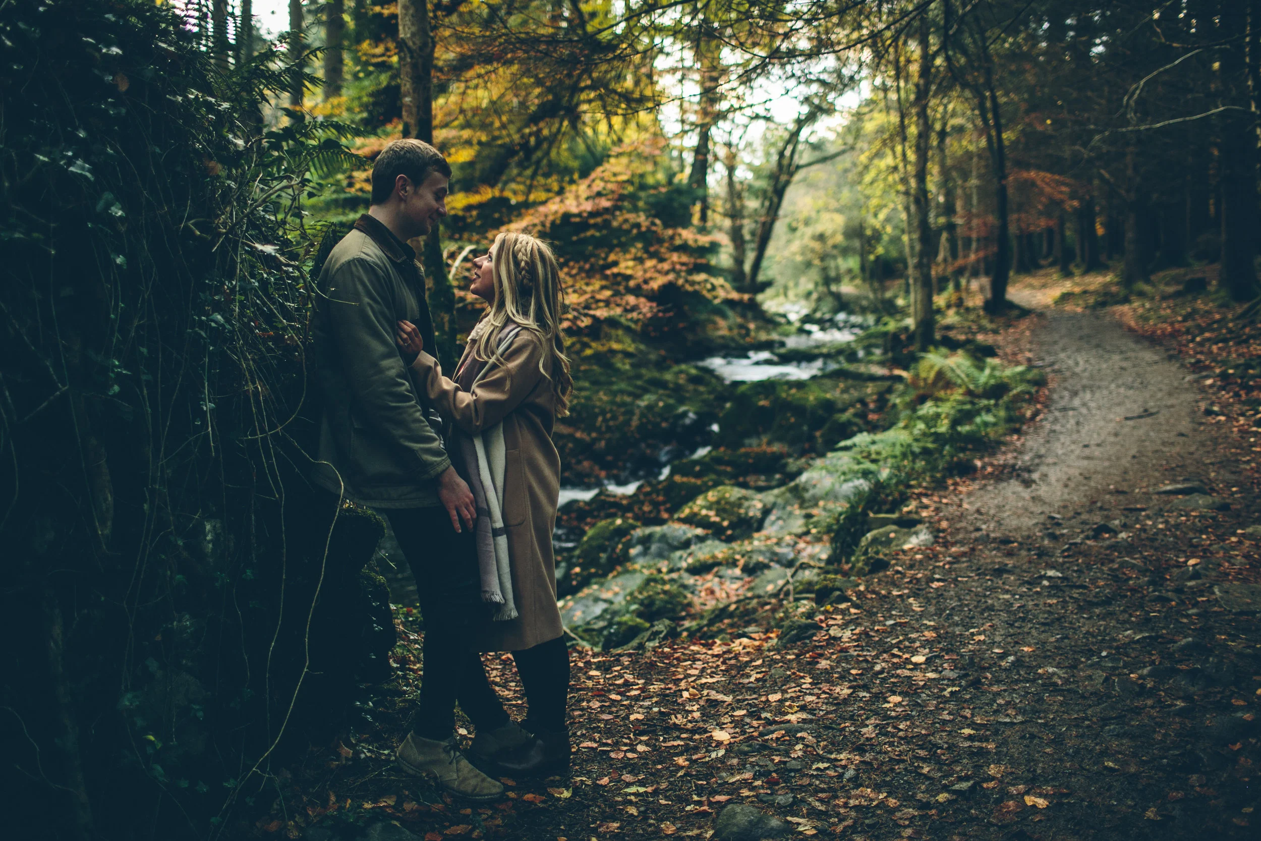 A man and woman stand close together on a wooded trail near a small stream in a forest during autumn, sharing an intimate moment.