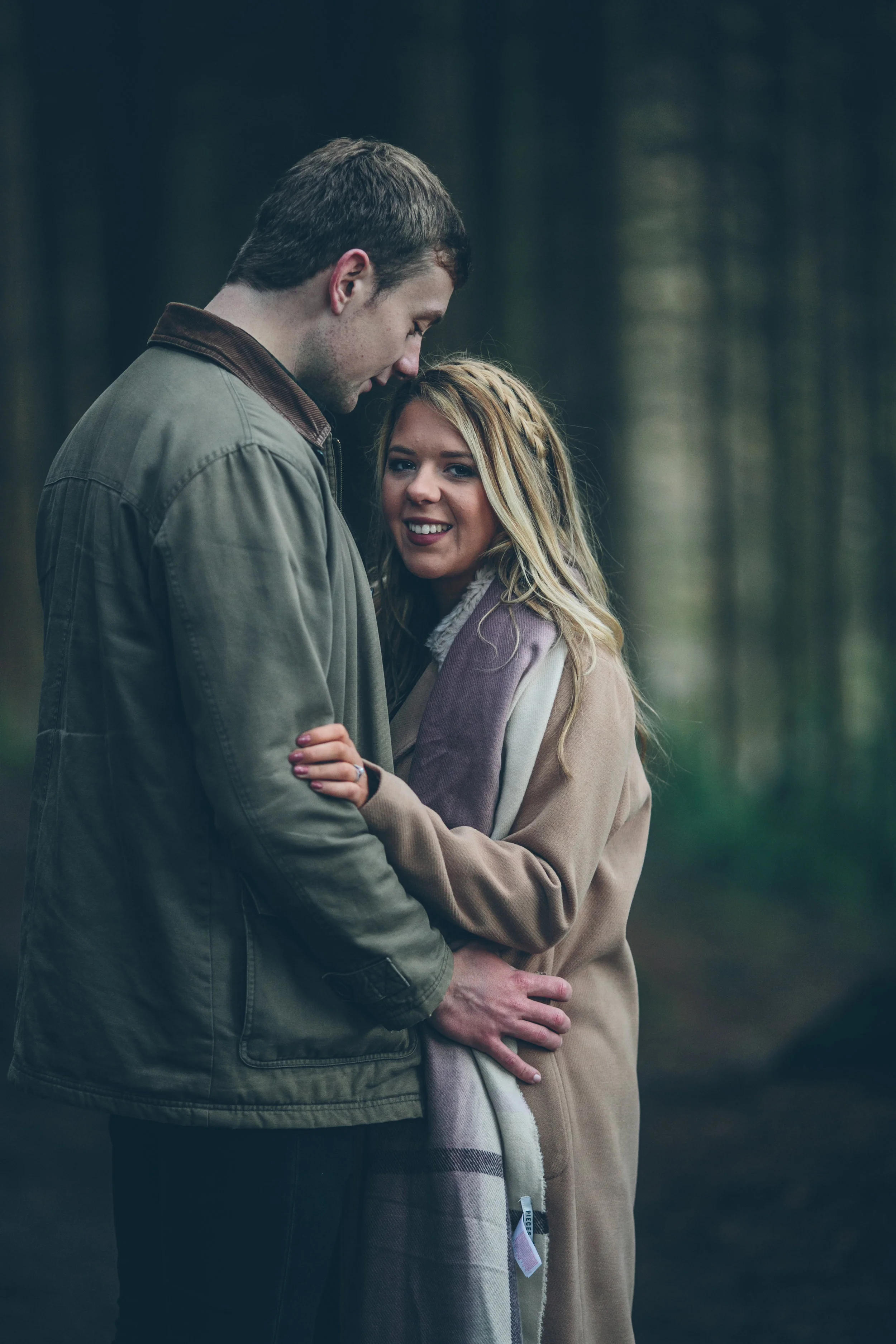 A young couple standing closely together in a forest, with the woman smiling and looking directly at the camera while the man looks down at her, both dressed in casual jackets.