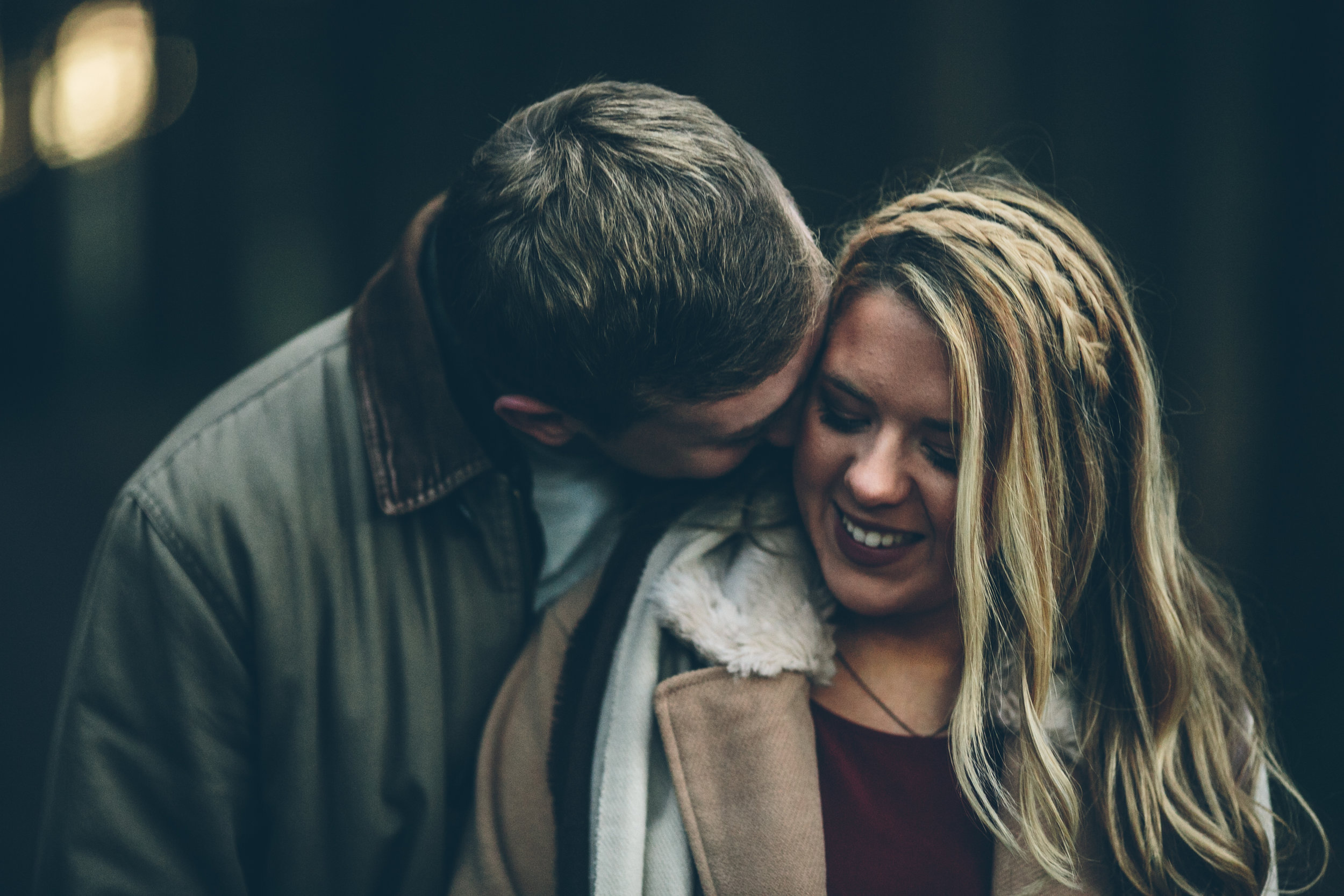 A man leaning in to kiss a woman on the cheek, both smiling, in a dark outdoor setting.