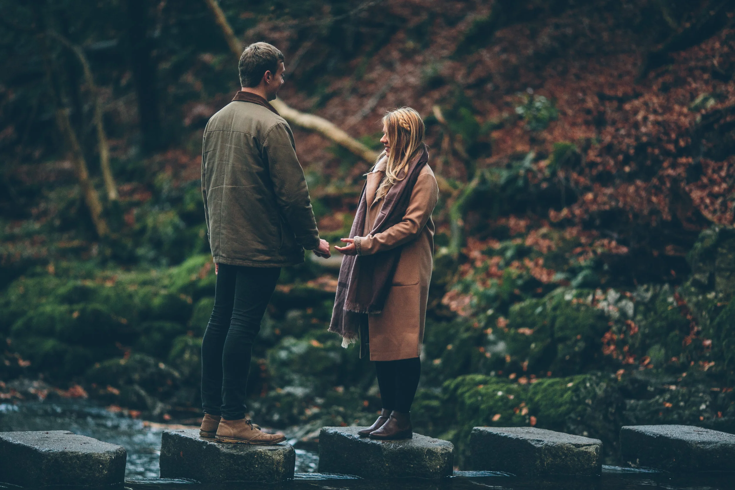 A couple is standing on stepping stones in a stream, holding hands and looking at each other, in a forest setting.