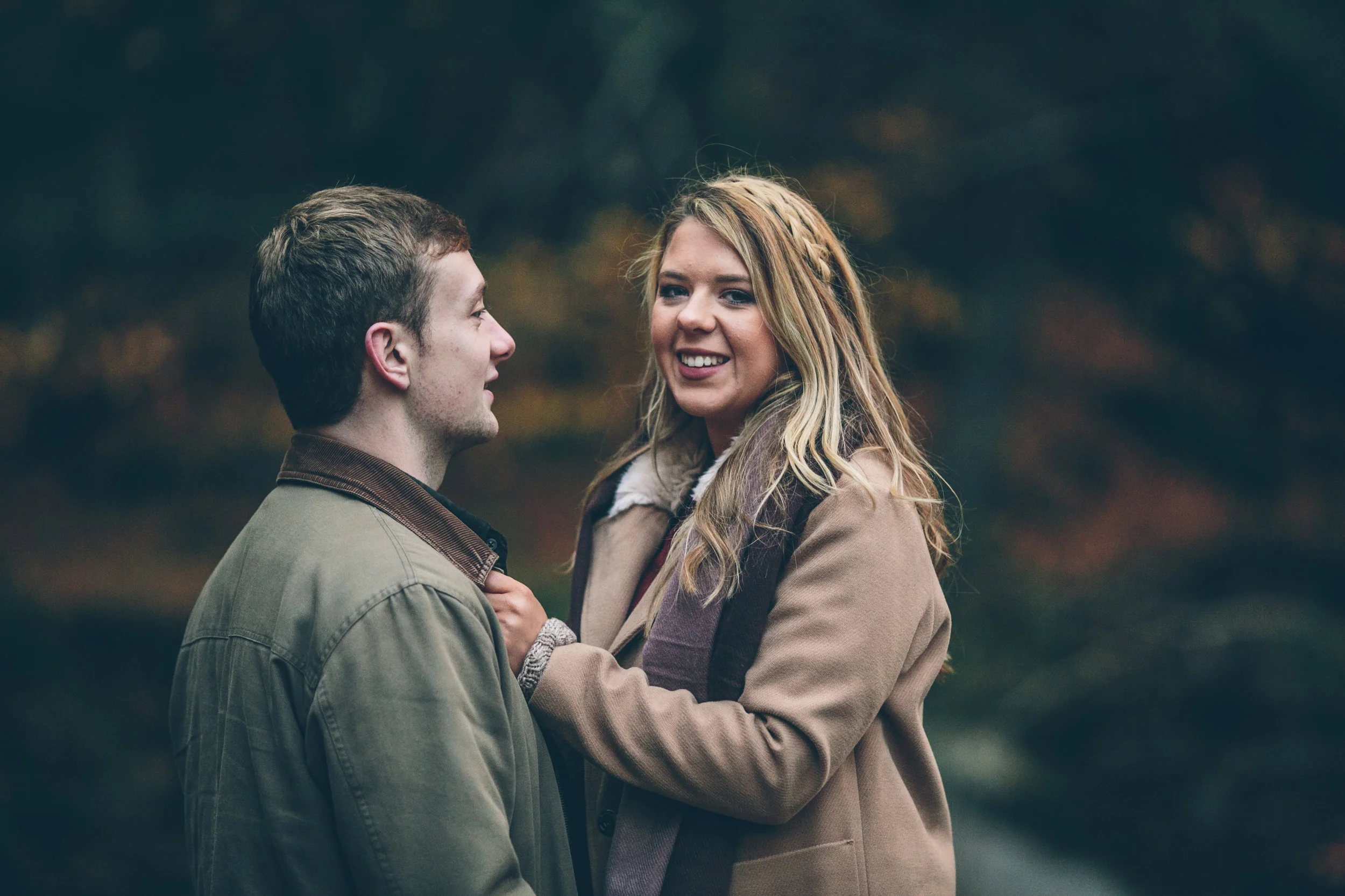 A man and woman outdoors in fall, facing each other closely with smiles, woman holding man's collar, blurred autumn background.
