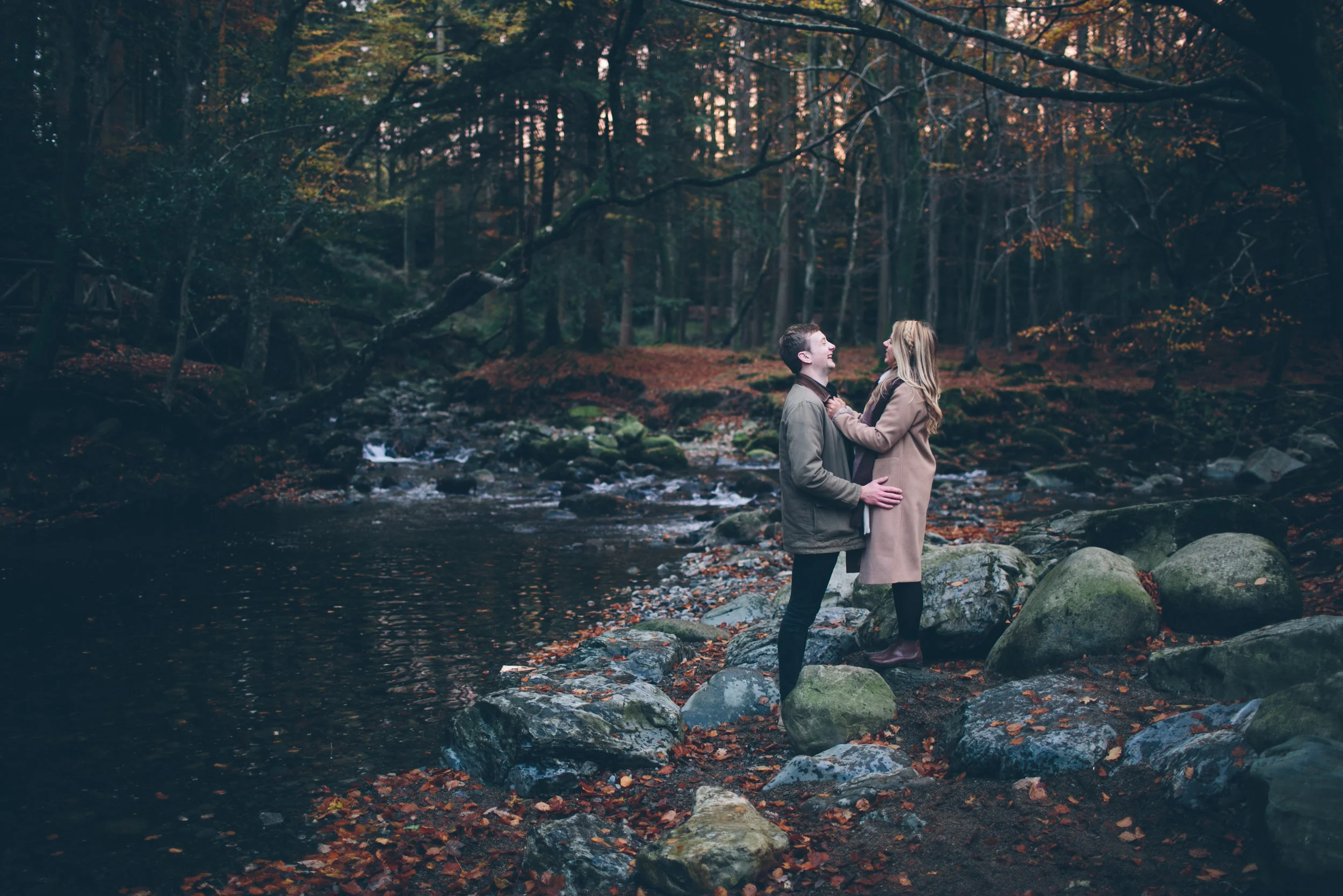 A couple stands on rocks by a river in a forest during autumn, smiling and looking into each other's eyes.