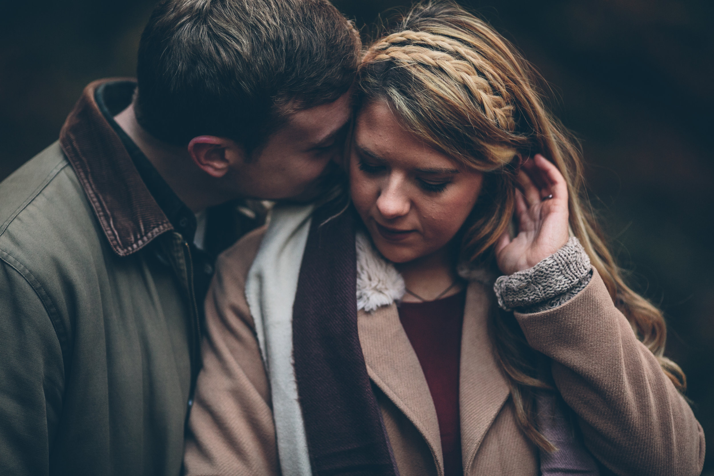 A man and a woman share a tender moment outdoors, with the man gently touching the woman's face as she looks down with a thoughtful expression.