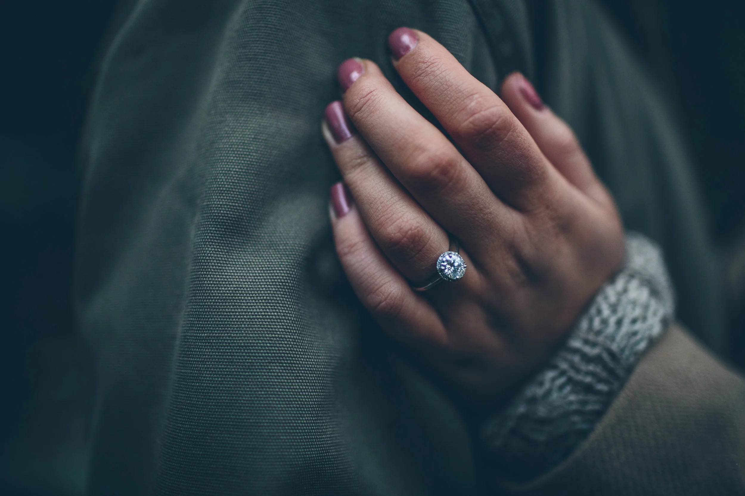 A woman resting her hand on her knee, showing a large diamond engagement ring on her finger, with pink nail polish and a knitted gray sleeve.