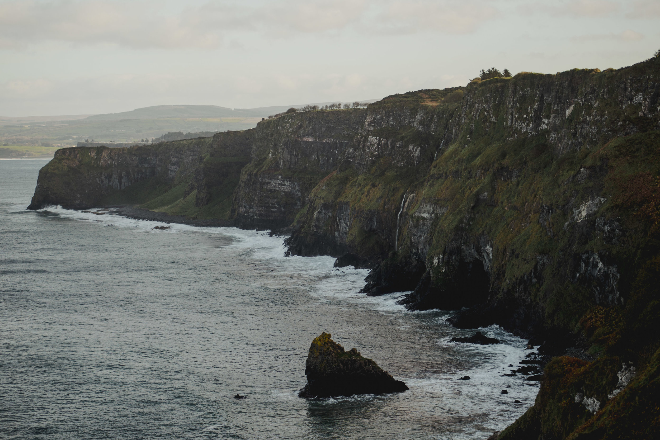 Cliffs along a dark ocean with waves crashing at the base and a small rocky outcrop in the water.