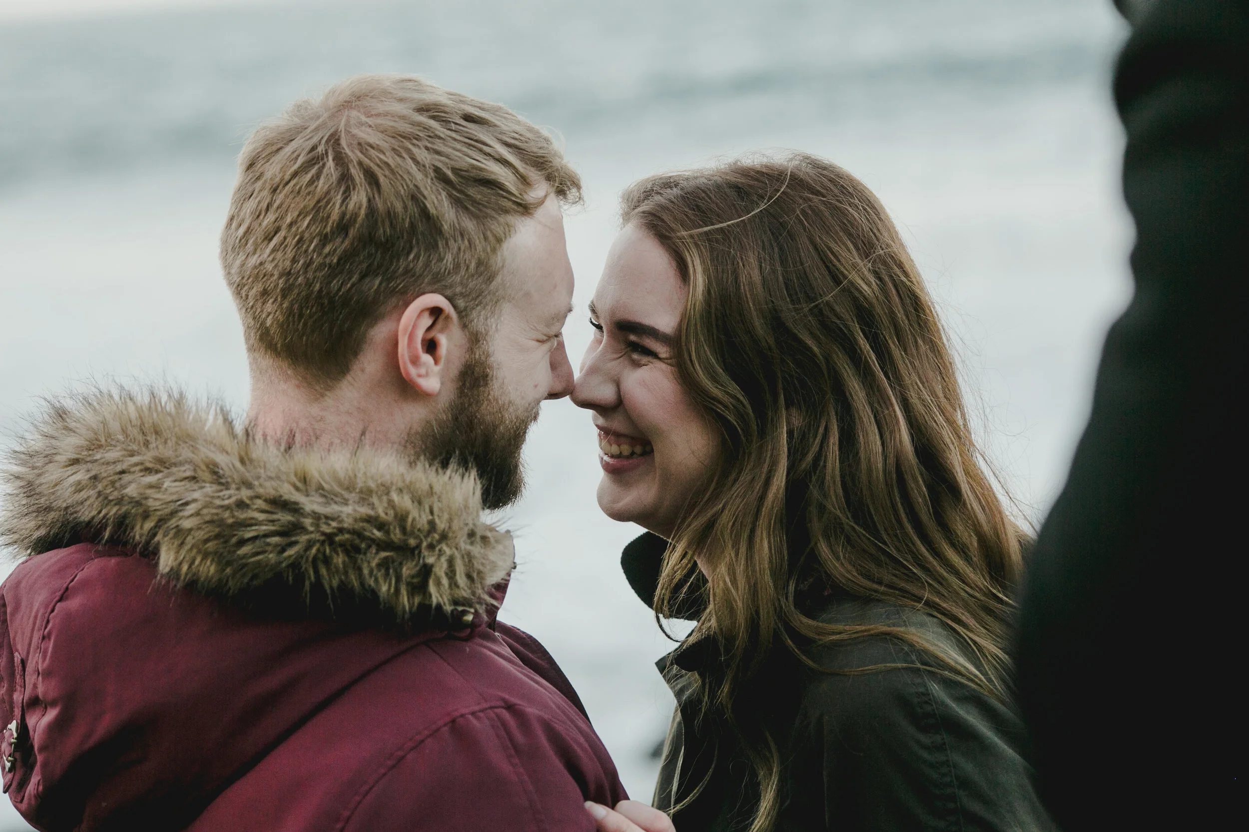 A couple with their foreheads touching and smiling at each other outdoors near the water, with a person partially visible on the right.