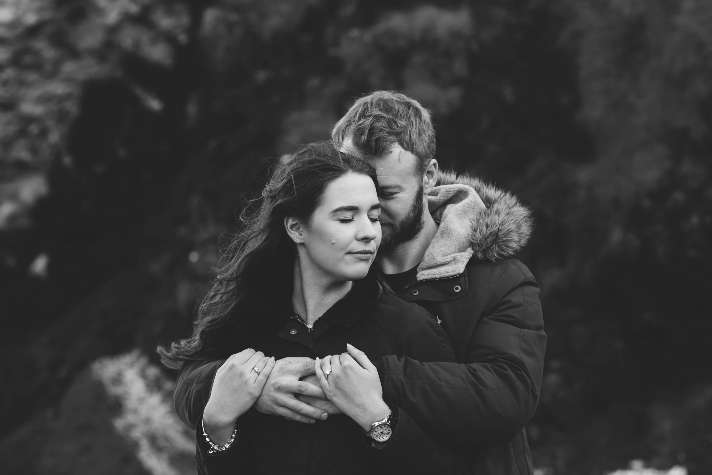 A black and white photo of a couple embracing outdoors, with the man gently touching the woman's shoulders and their eyes closed, conveying tenderness and affection.