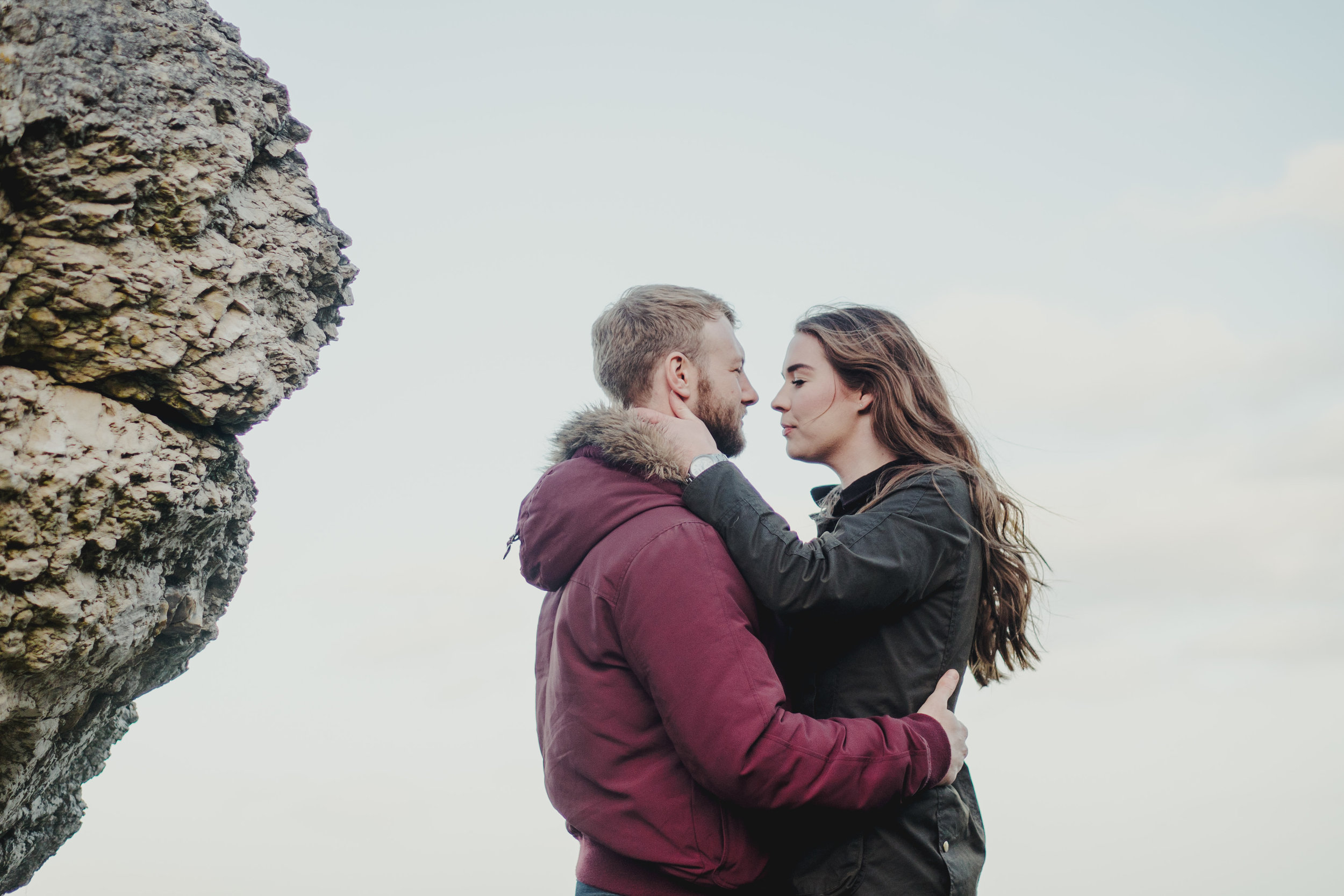 A couple facing each other outdoors, with the woman holding the man’s face and the man holding her waist, near a large rock formation, against a cloudy sky.