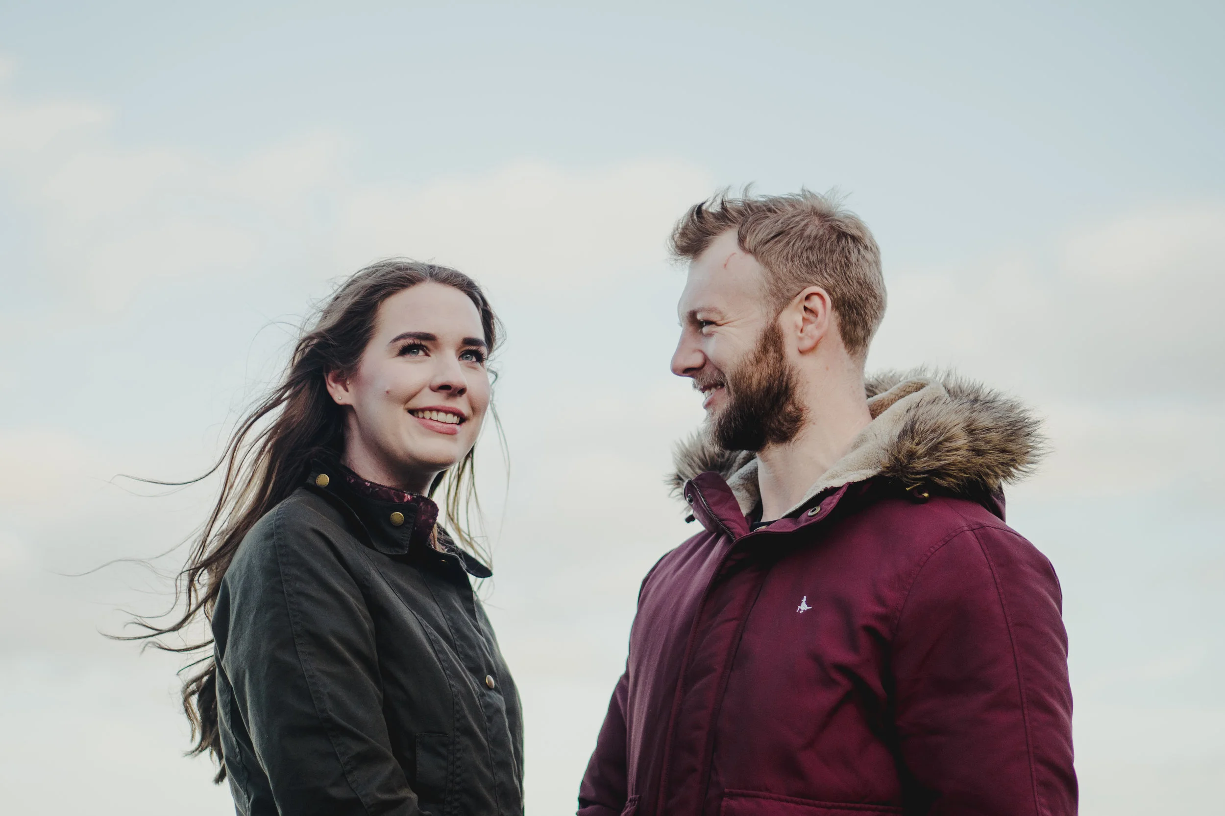 A smiling woman and man facing each other outdoors against a cloudy sky, wearing warm jackets. Couples Session. Kinbane Castle.