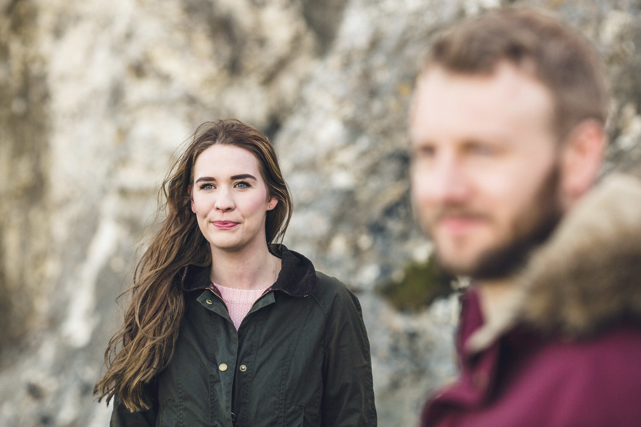 A woman with long brown hair and blue eyes looking at the camera, wearing a black jacket with a pink shirt underneath, standing outdoors with a blurred man in the foreground and rocky background. Couples Session. Kinbane Castle.