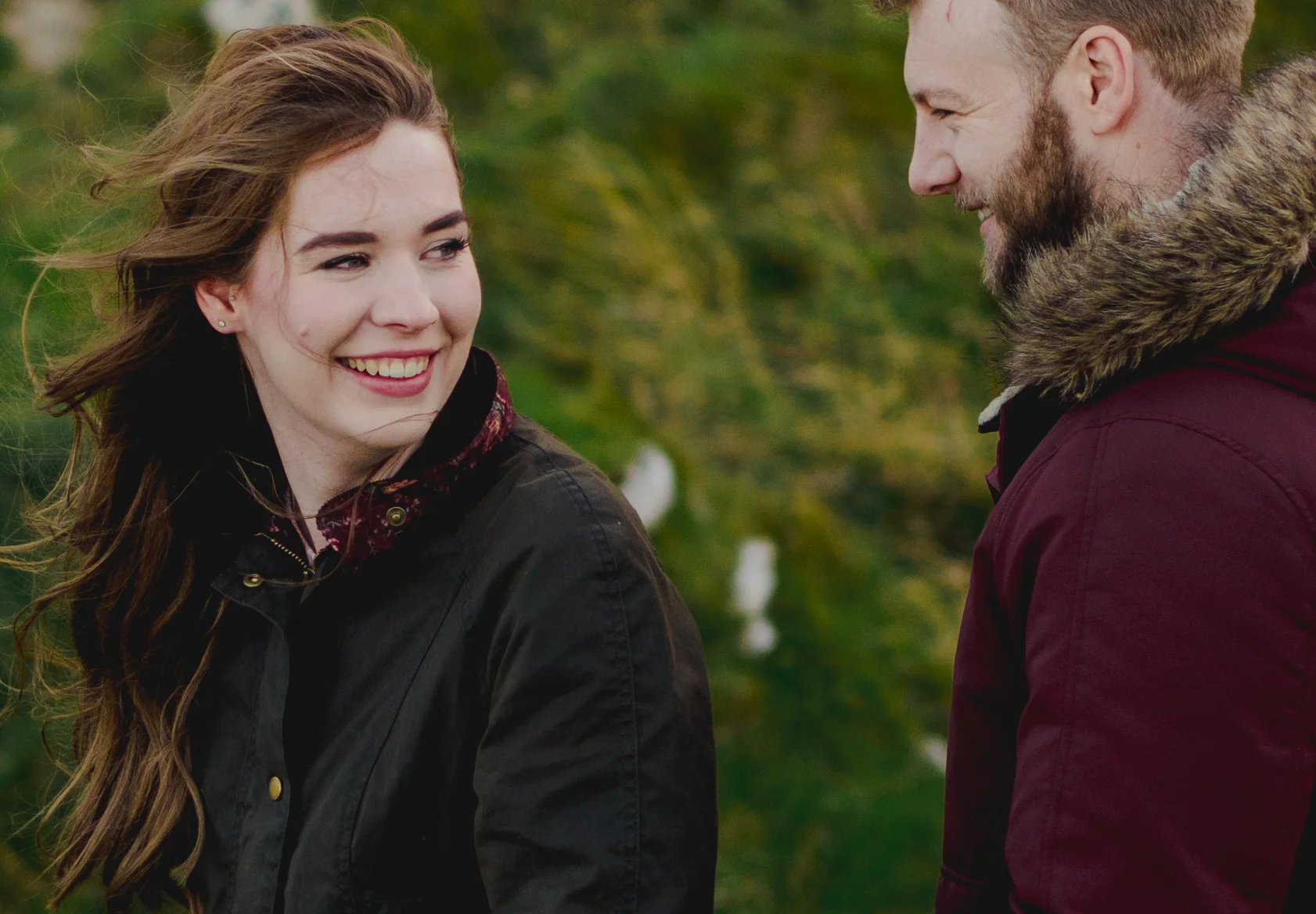 A woman with long brown hair smiling and looking at a man with a beard. They are outdoors with blurred green trees in the background. Both wear jackets, the woman's dark and the man's maroon with a fur-lined hood.