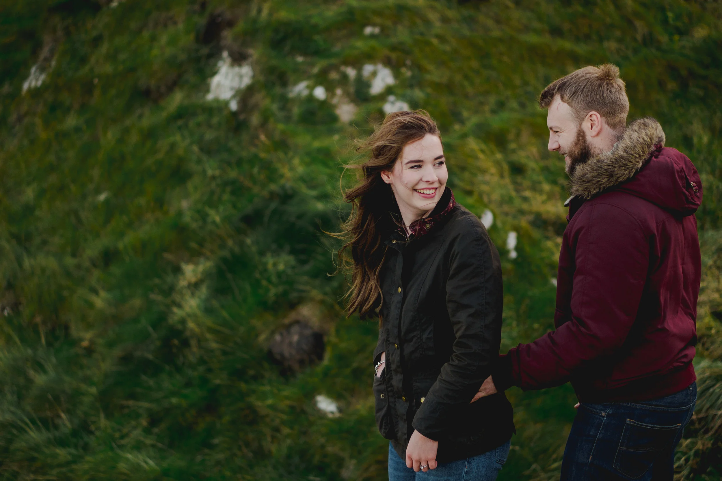 A young woman and a young man outdoors, smiling and laughing together. The woman is wearing a black jacket and the man is wearing a maroon jacket with a fur-lined hood. They are holding hands and facing each other in a lush green, hilly landscape.