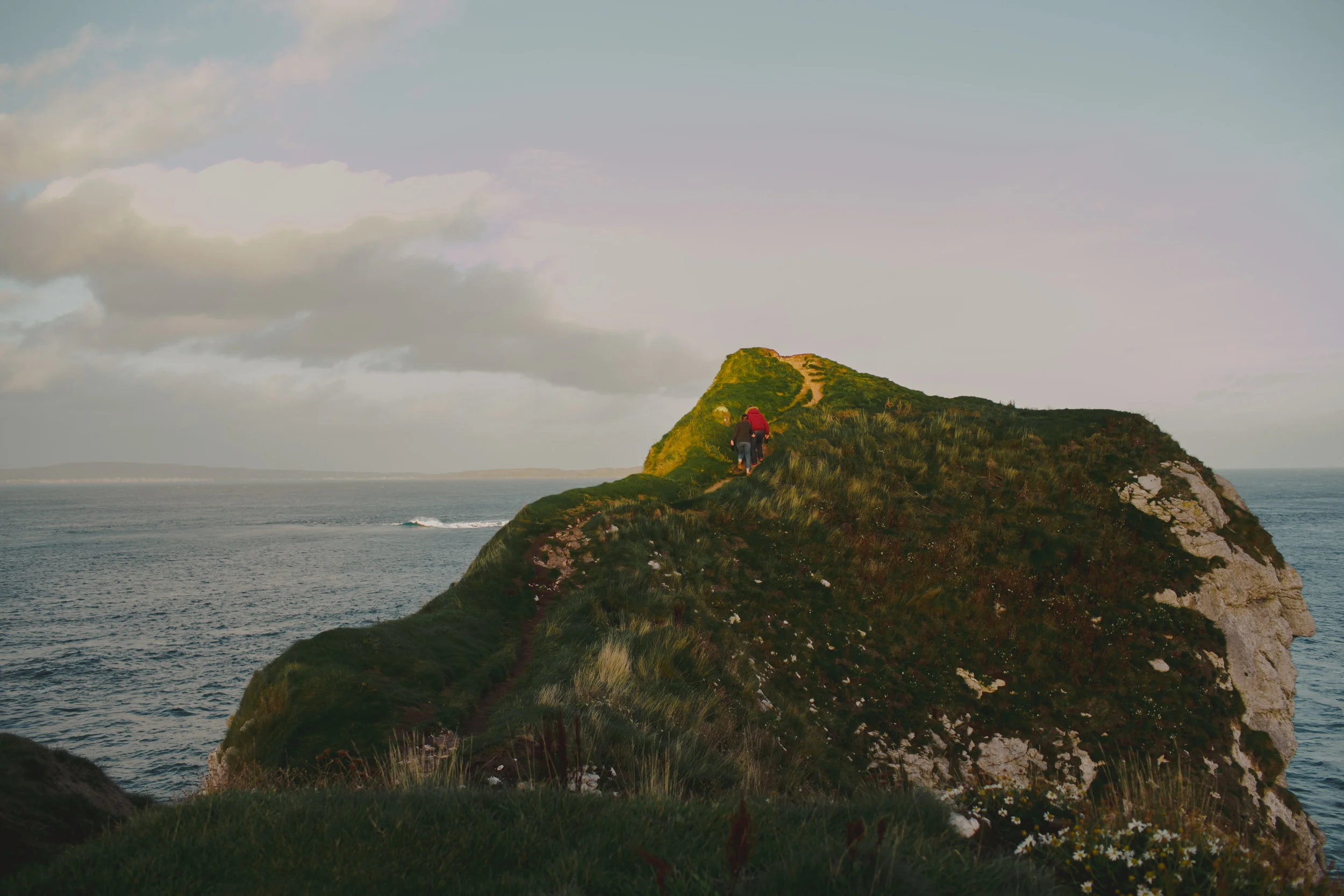 Two hikers walking along a grassy, narrow trail on a hill overlooking the ocean, with a cloudy sky overhead. Couples Session. Kinbane Castle.