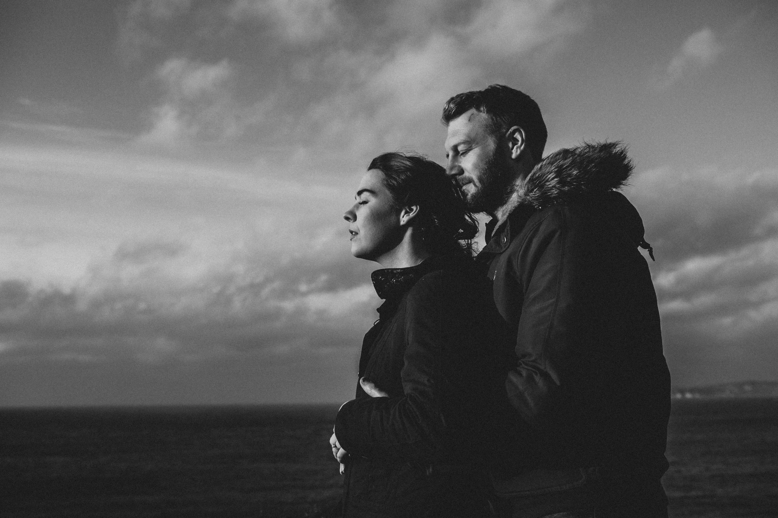 A man and woman standing outdoors against a cloudy sky, embracing each other with eyes closed, black and white photo.