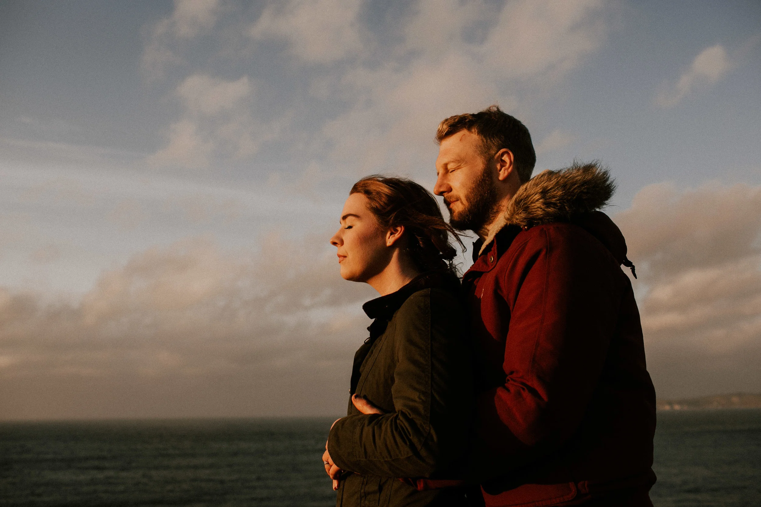 A couple stands close together outdoors near the ocean, with eyes closed, embracing as they enjoy the peaceful moment at sunset.