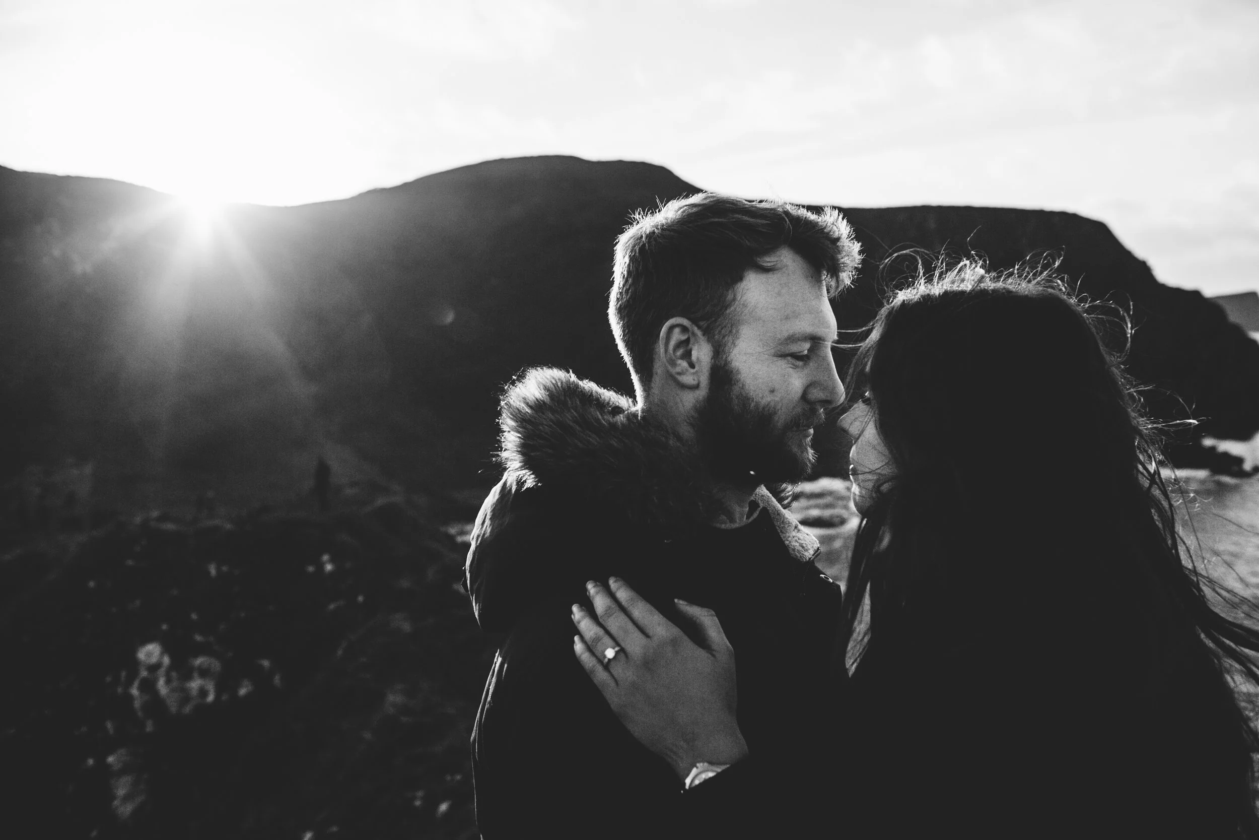 A black and white photo of a man and woman embracing outdoors at sunset with mountains in the background.