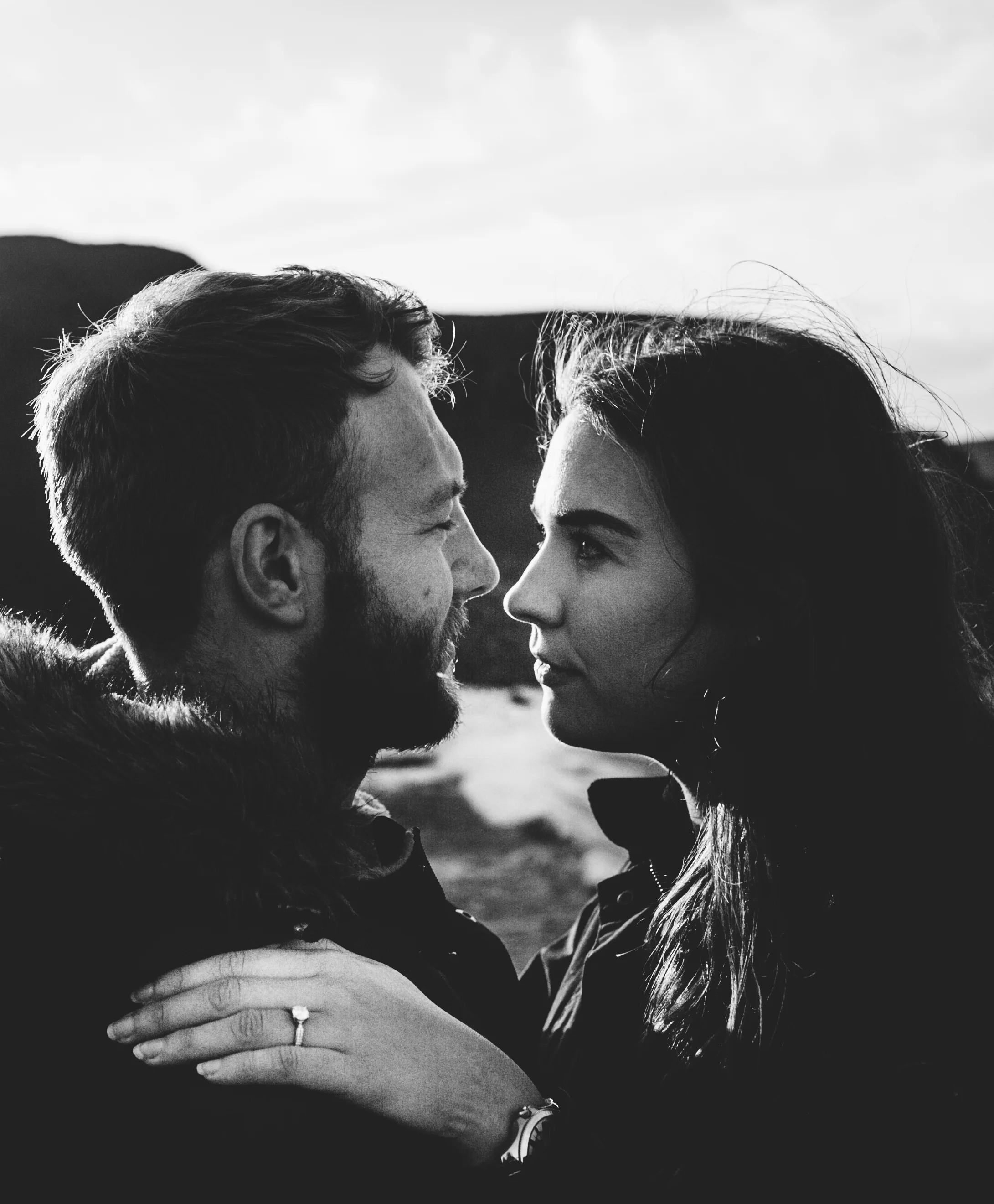 A black and white photo of a man and woman close together outdoors, looking into each other's eyes, with a landscape background.
