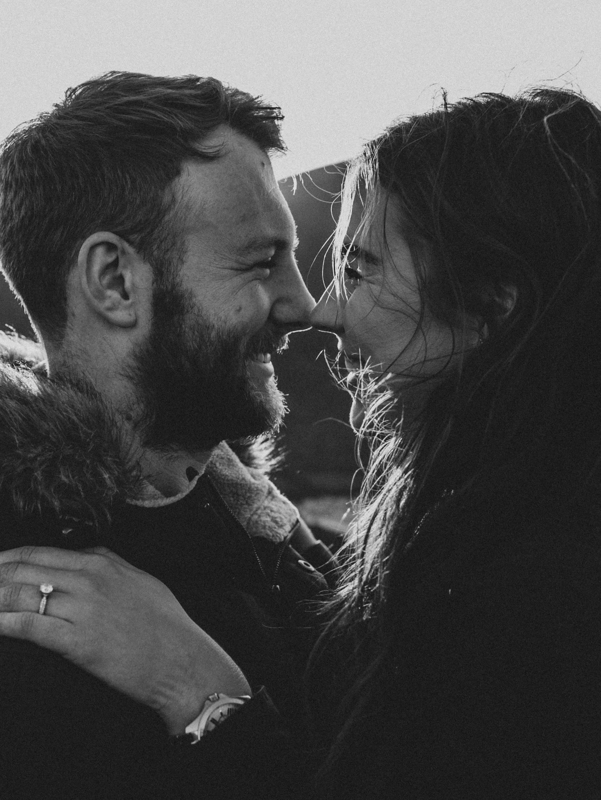 A black-and-white photo of a couple smiling and touching noses, close-up shot. The man has a beard and is wearing a coat with a fur-lined collar, and the woman has long hair and is wearing a dark top. The woman's hand with an engagement ring rests on