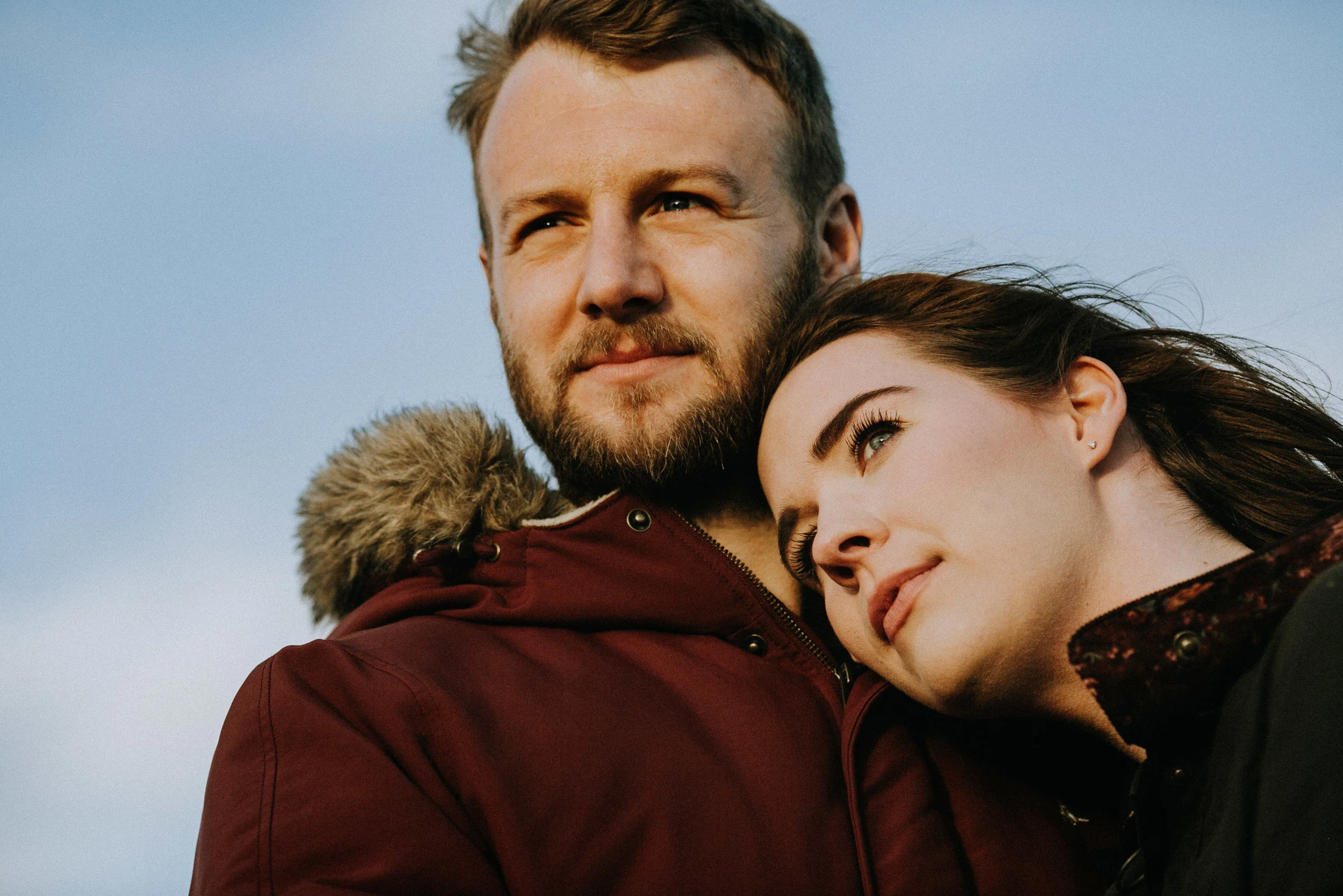 A man with a beard and blonde hair, and a woman with dark hair, are close together outdoors against a blue sky. The woman is resting her head on the man's shoulder, and both are looking content.