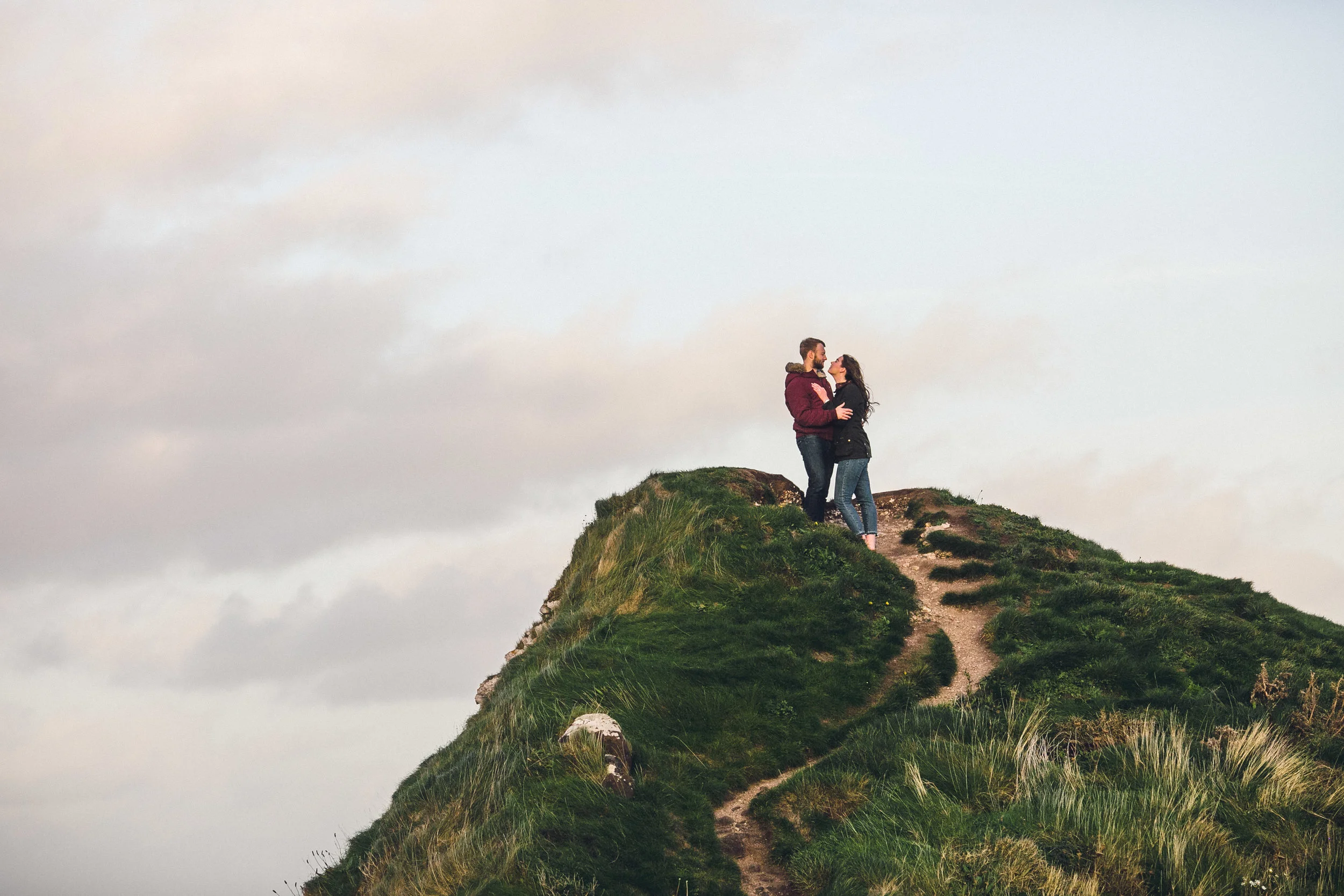 A couple stands on the top of a grassy hill, embracing and looking at each other, with a cloudy sky in the background. Couples Session. Kinbane Castle.