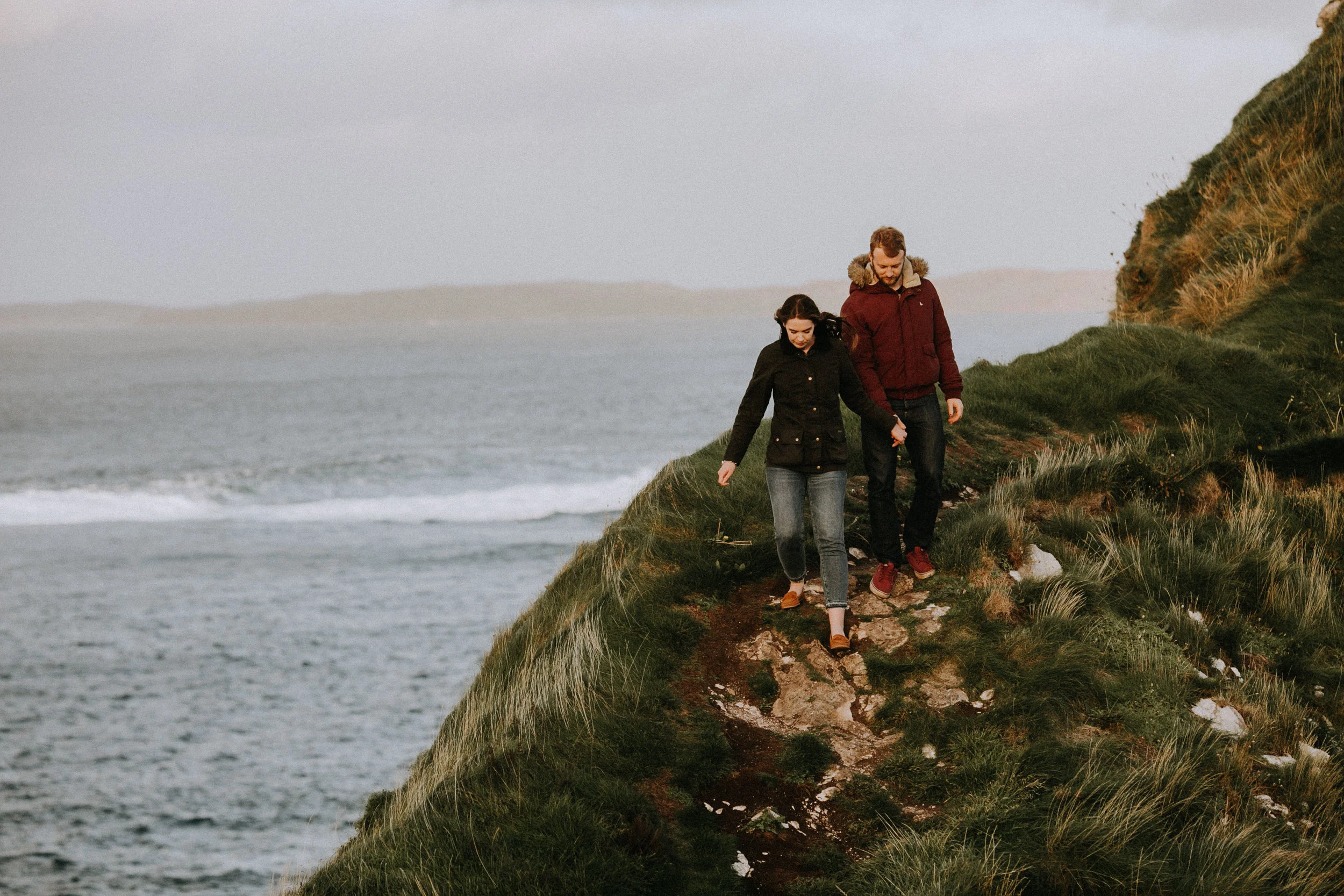 A couple walking hand in hand along a grassy coastal cliff path with the ocean in the background.