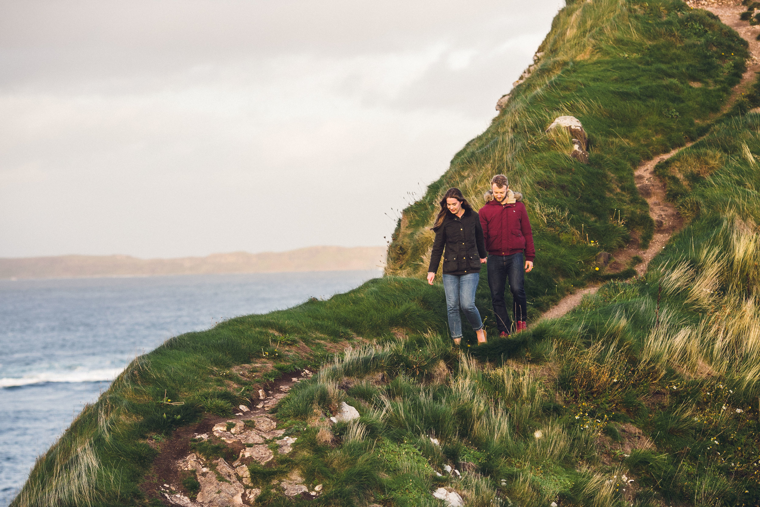 A man and a woman walk along a narrow, grassy trail on a coastal hillside with ocean in the background.