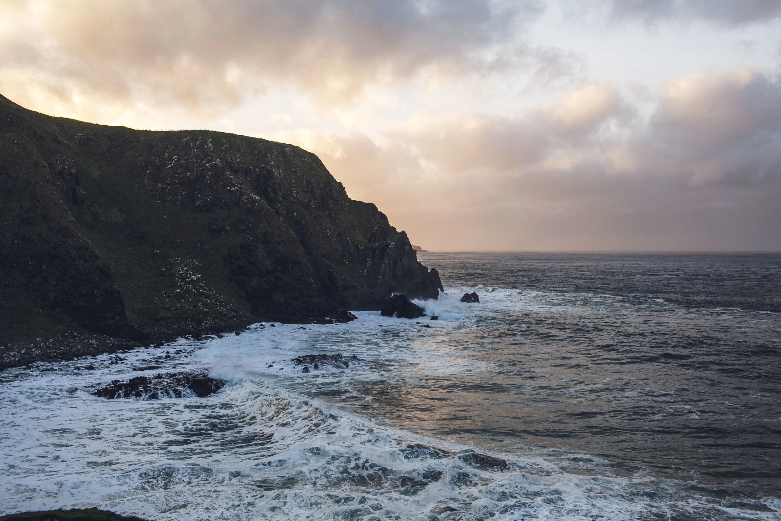 A rugged coastline with steep green cliffs meeting the ocean, waves crashing at the base of the cliffs, under a cloudy sky at sunset or sunrise.