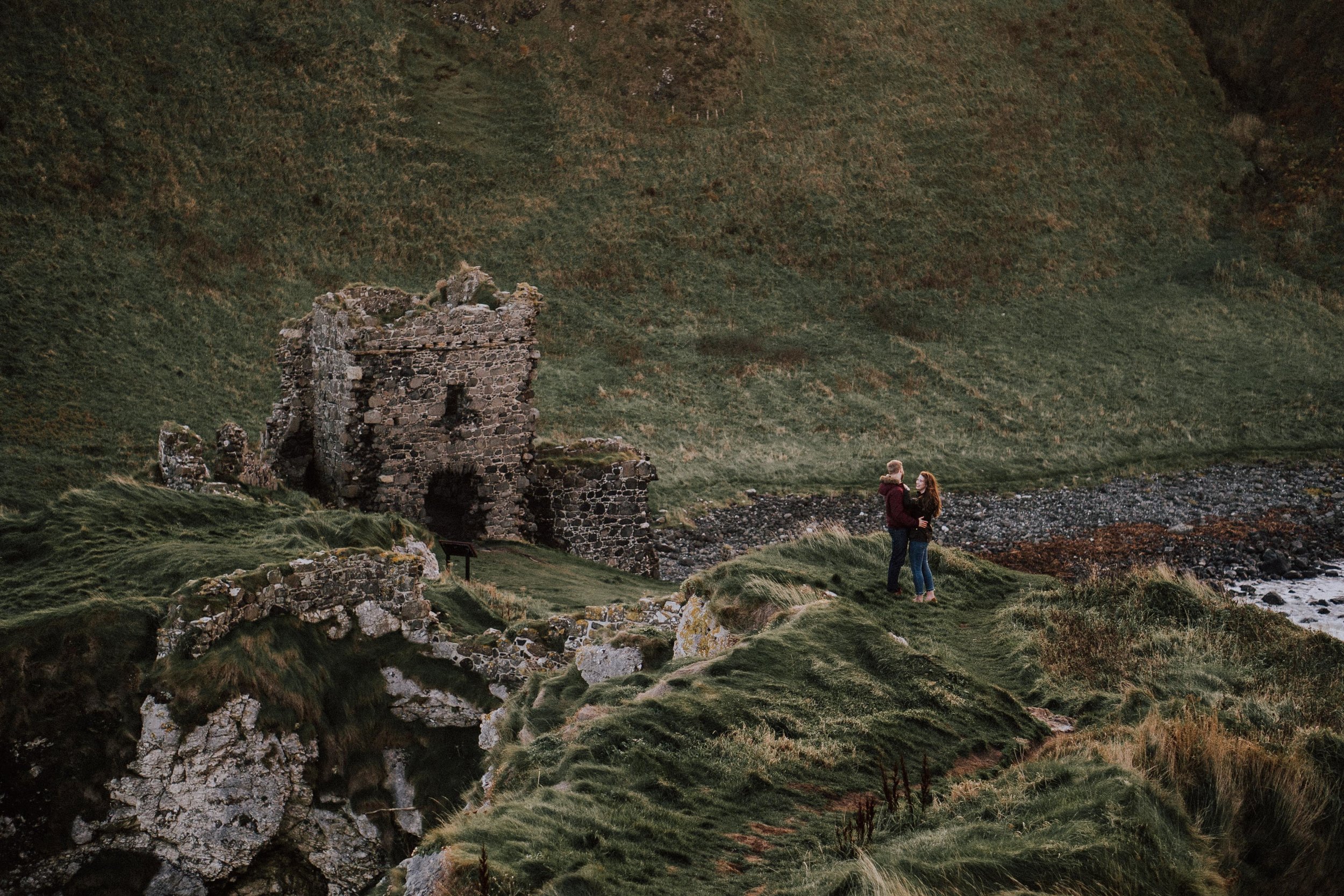 Two people standing on green grassy landscape near an old stone castle ruin, with hills and a river in the background.